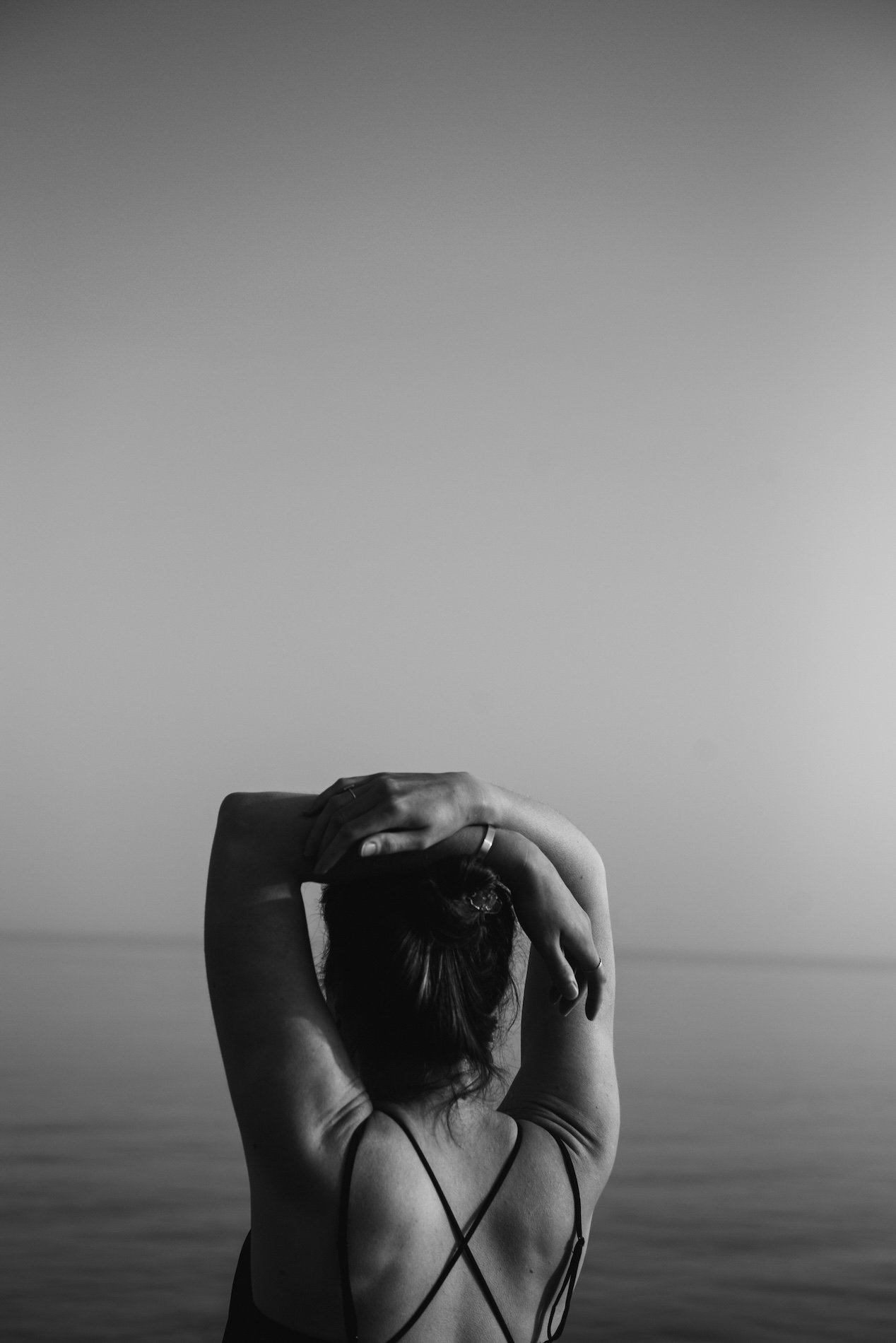 A woman standing with her back facing the camera, arms raised and hands resting on her head, near a body of water with a clear sky in the background in black and white.