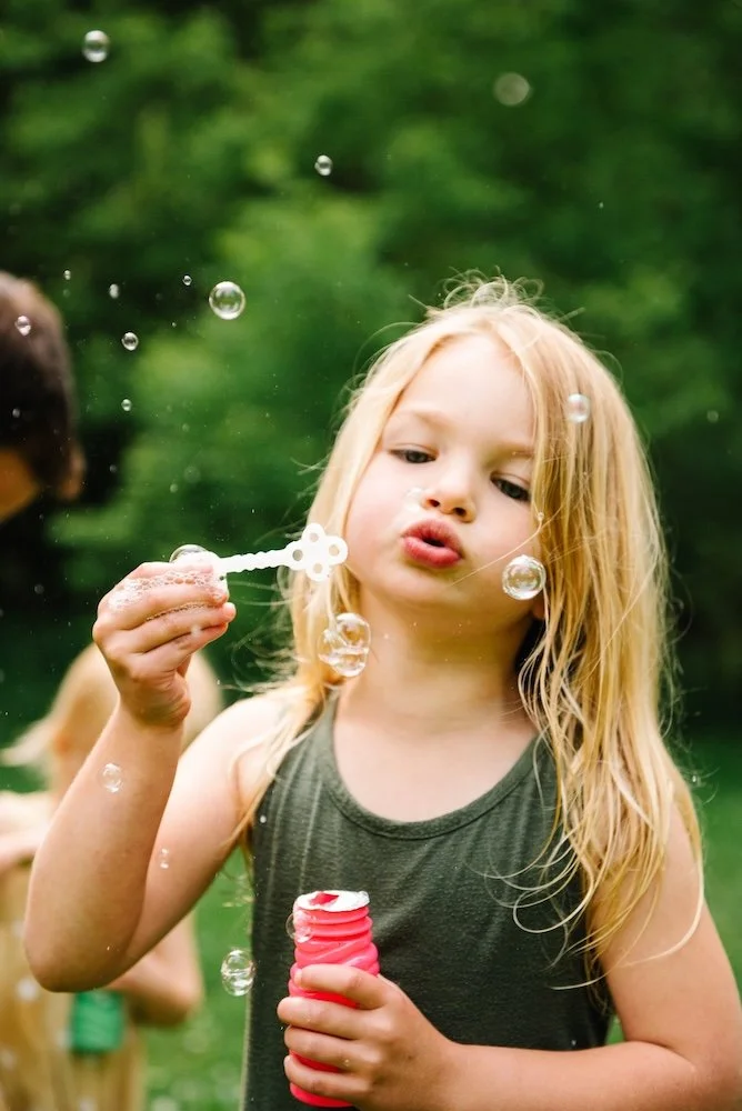 A young girl with blonde hair blowing bubbles outdoors, holding a bubble wand and a bottle of bubble solution, with a green blurred background.