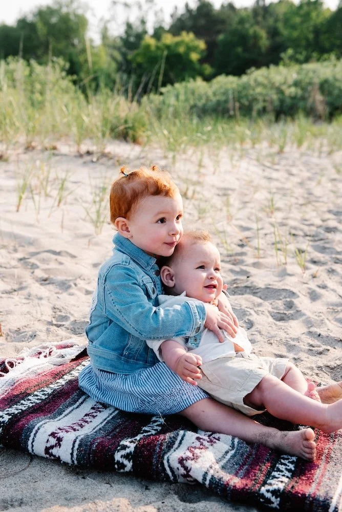 Two young children sitting on a blanket on a sandy beach, with greenery and trees in the background, during daylight.