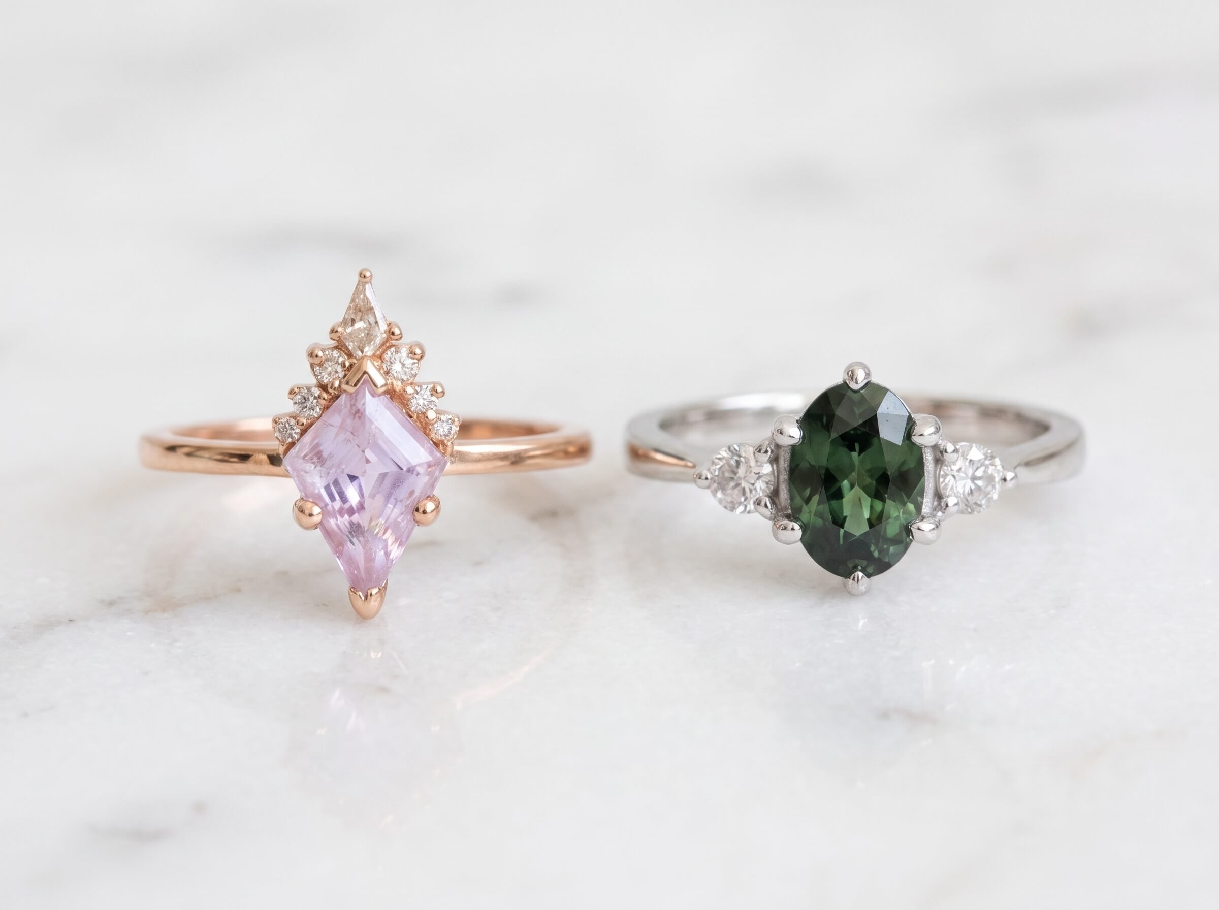 Two rings displayed on marble - a rose gold ring with a kite-shaped lavender gemstone and diamond crown cluster, and a white gold three-stone ring with an oval deep green center gemstone and round diamond sides.