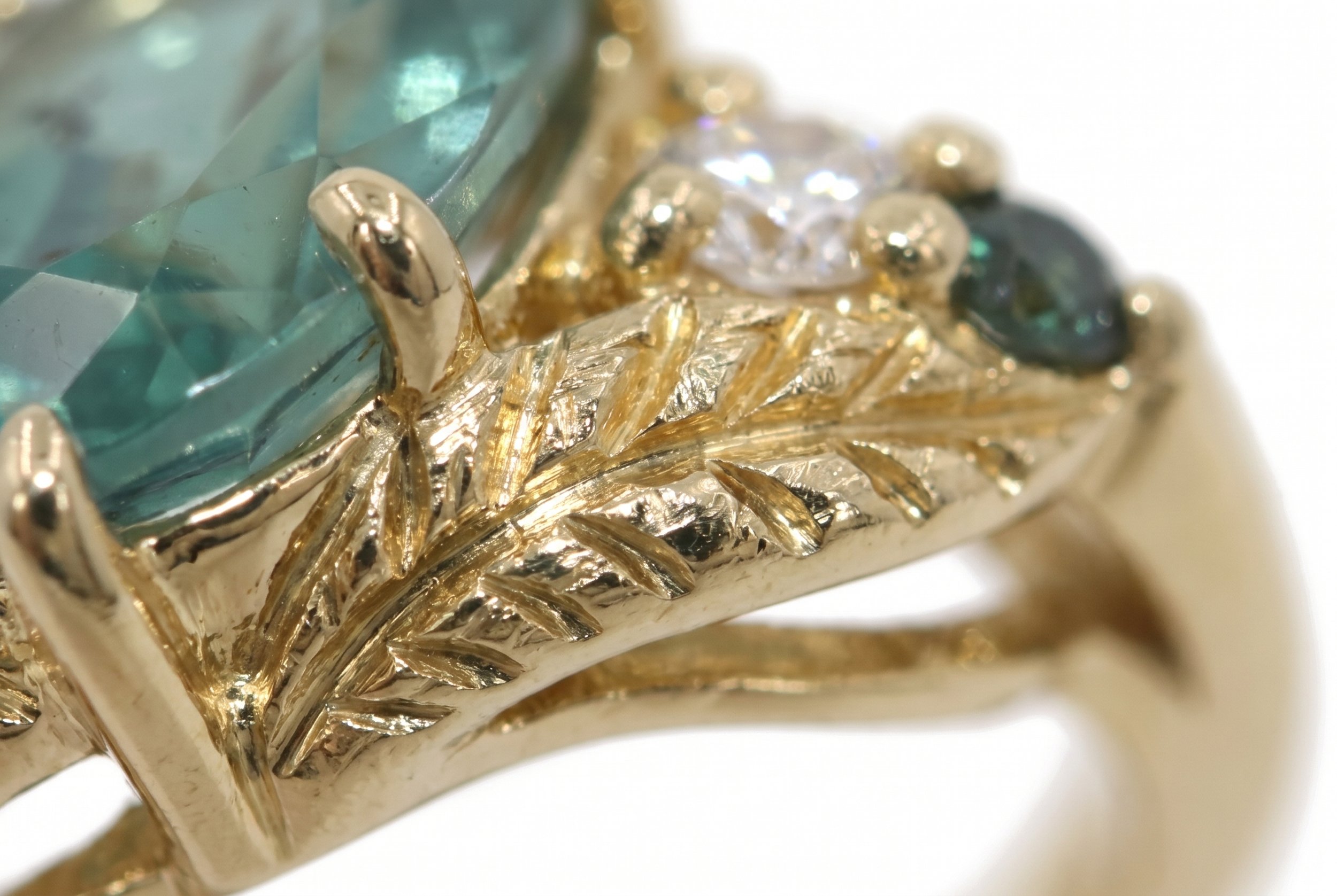An extreme close-up of hand-engraved leaf and branch detailing on a yellow gold ring setting, flanked by a round diamond and a small dark teal accent stone.