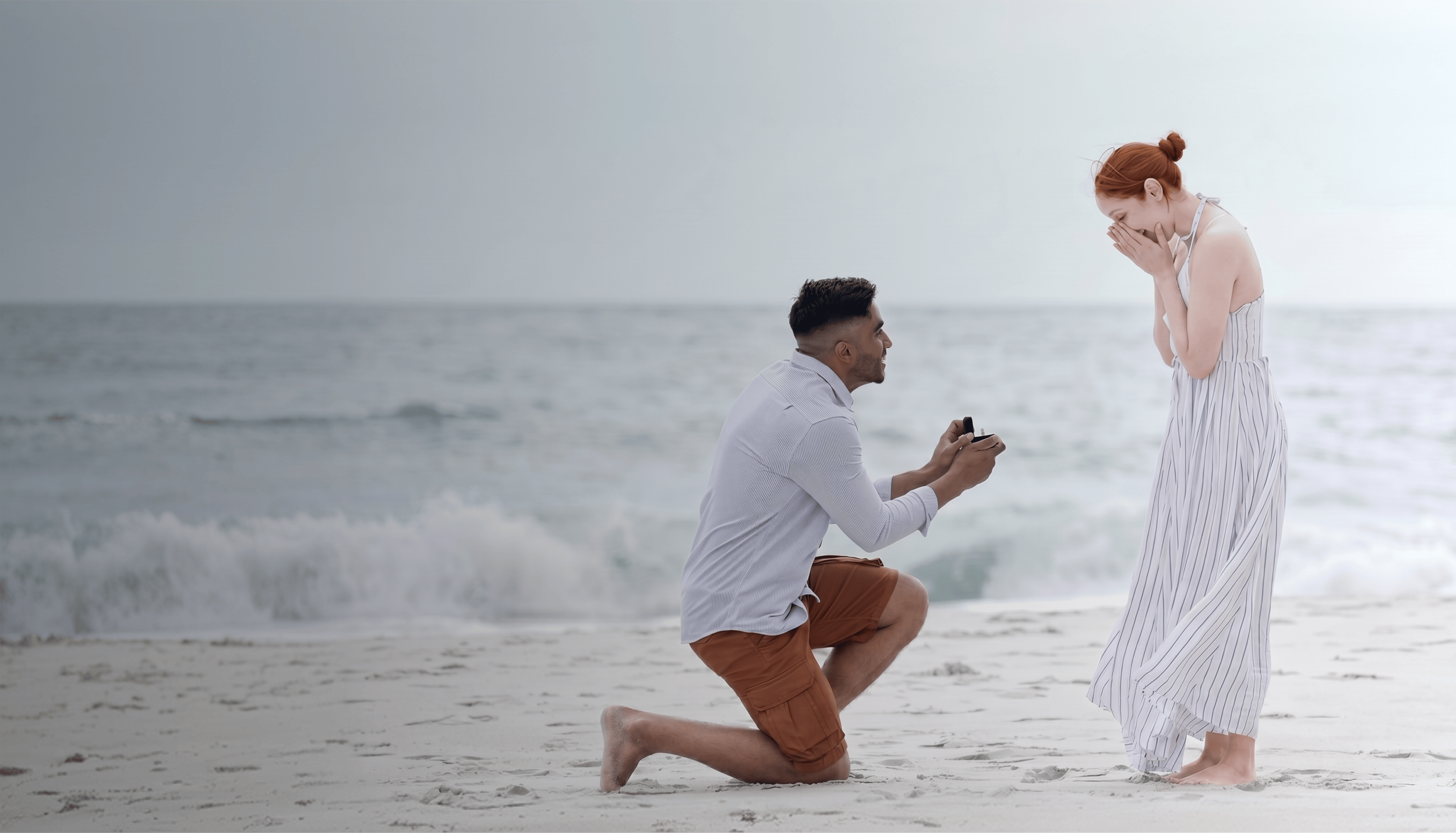 A man kneeling on the beach proposing to a woman who is covering her mouth in surprise.