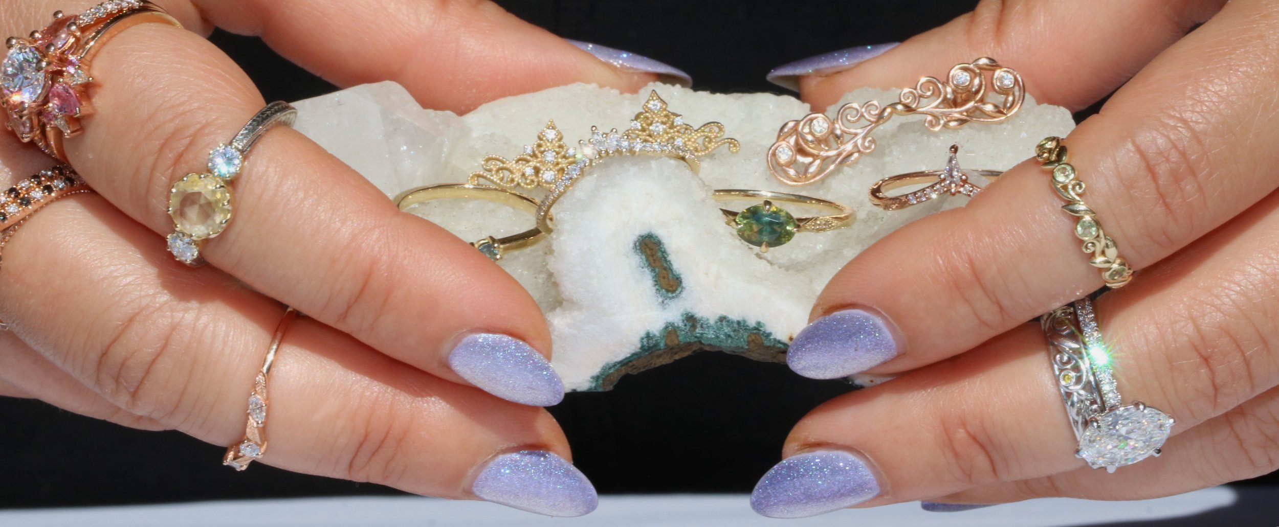 Close-up of hands holding a piece of white stone with rings and jewelry on fingers, including gold, silver, and rose gold rings with various gemstones and intricate designs.