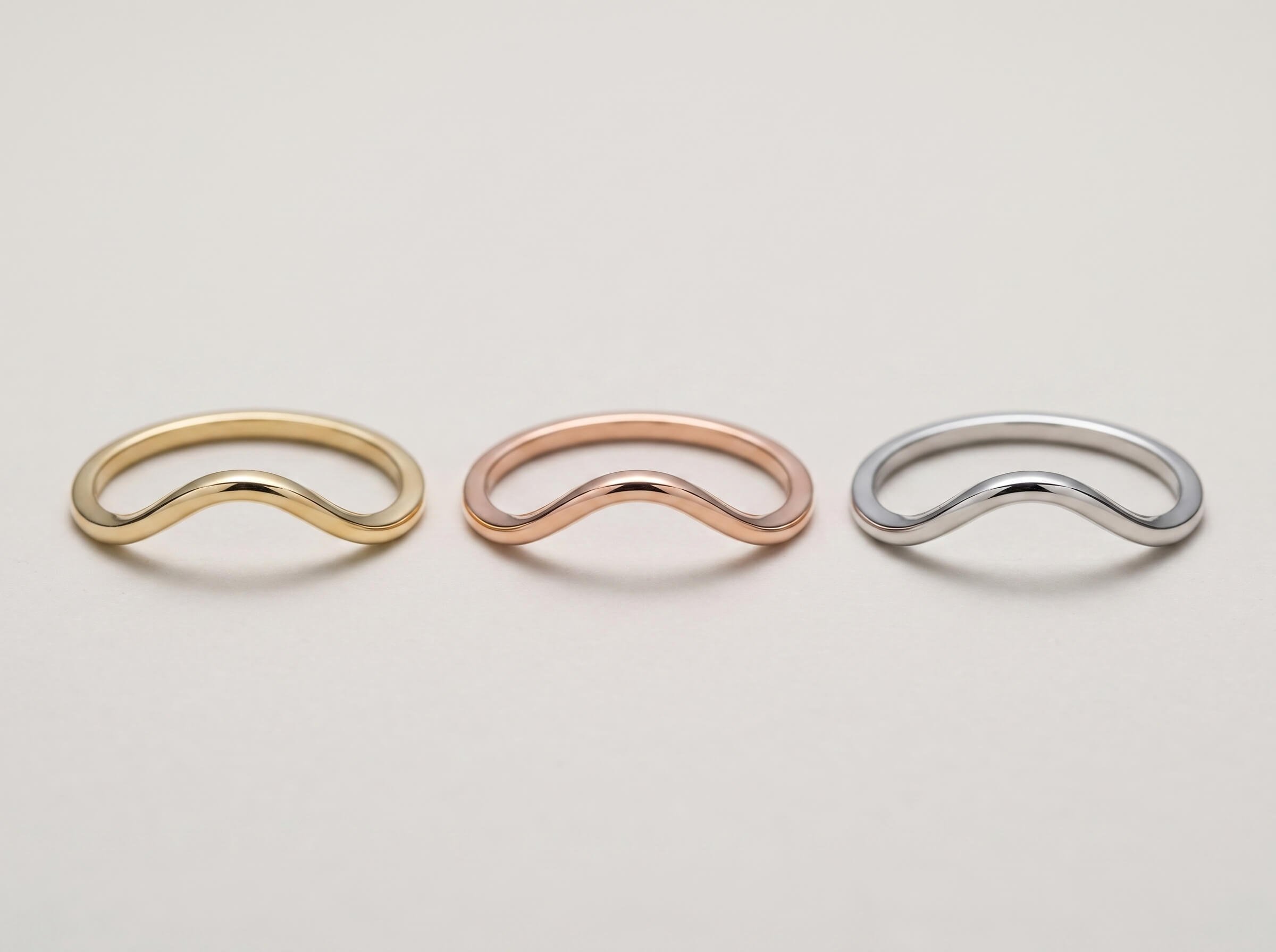 A flat-lay of four custom rings on linen - a green gemstone three-stone ring in yellow gold, a round diamond with pink halo, a yellow gold filigree band with ruby accents, and a wide matte white gold band.