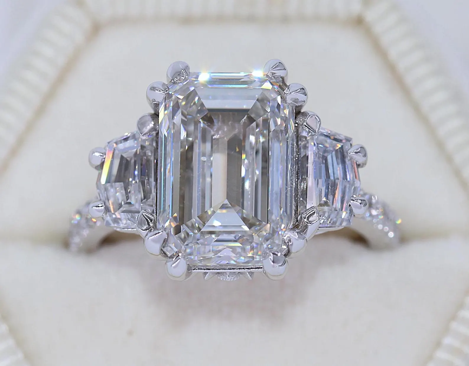 A large, emerald-cut diamond ring with smaller diamonds surrounding the center stone, set in a silver band, inside a white ring box