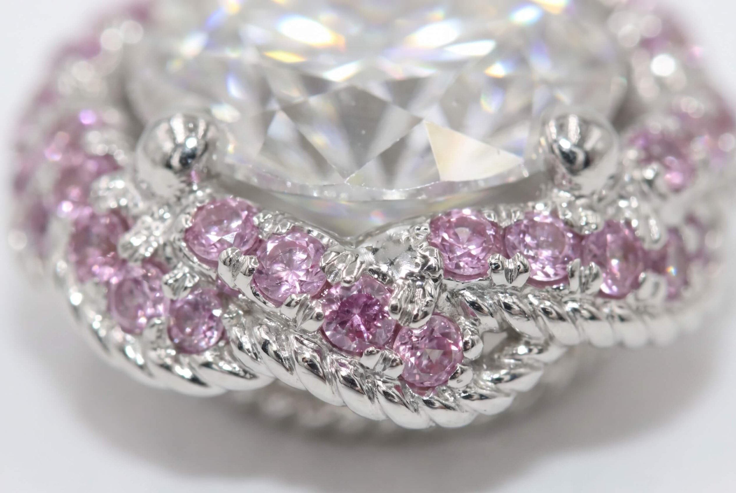 An extreme close-up of a pink sapphire halo setting, showing two rows of round pink stones set between twisted rope-style white gold detailing beneath a large round diamond.