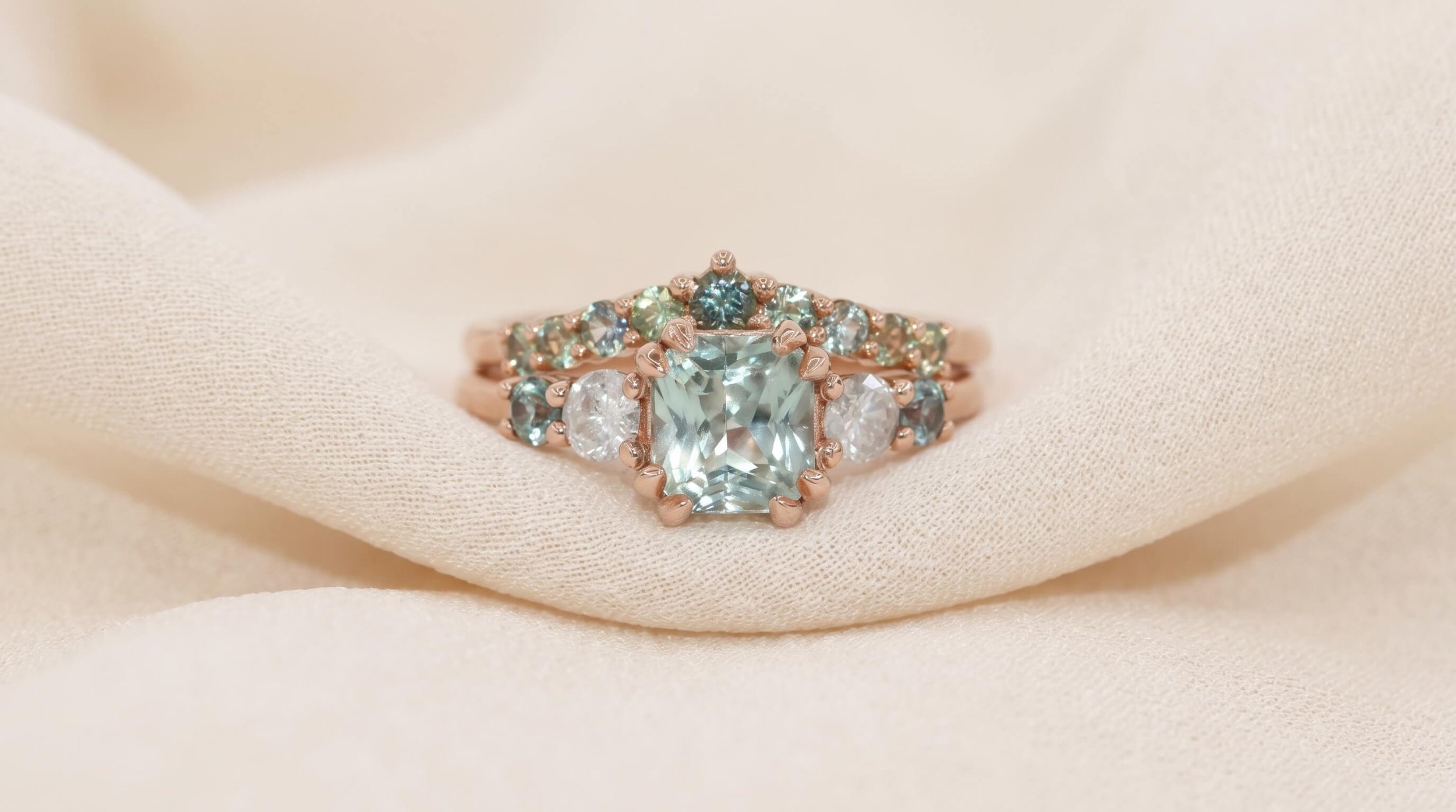 A rose gold ring set with a radiant-cut mint green gemstone, flanked by round diamonds and a curved wedding band with graduating teal and green sapphire accents, displayed on draped cream fabric.