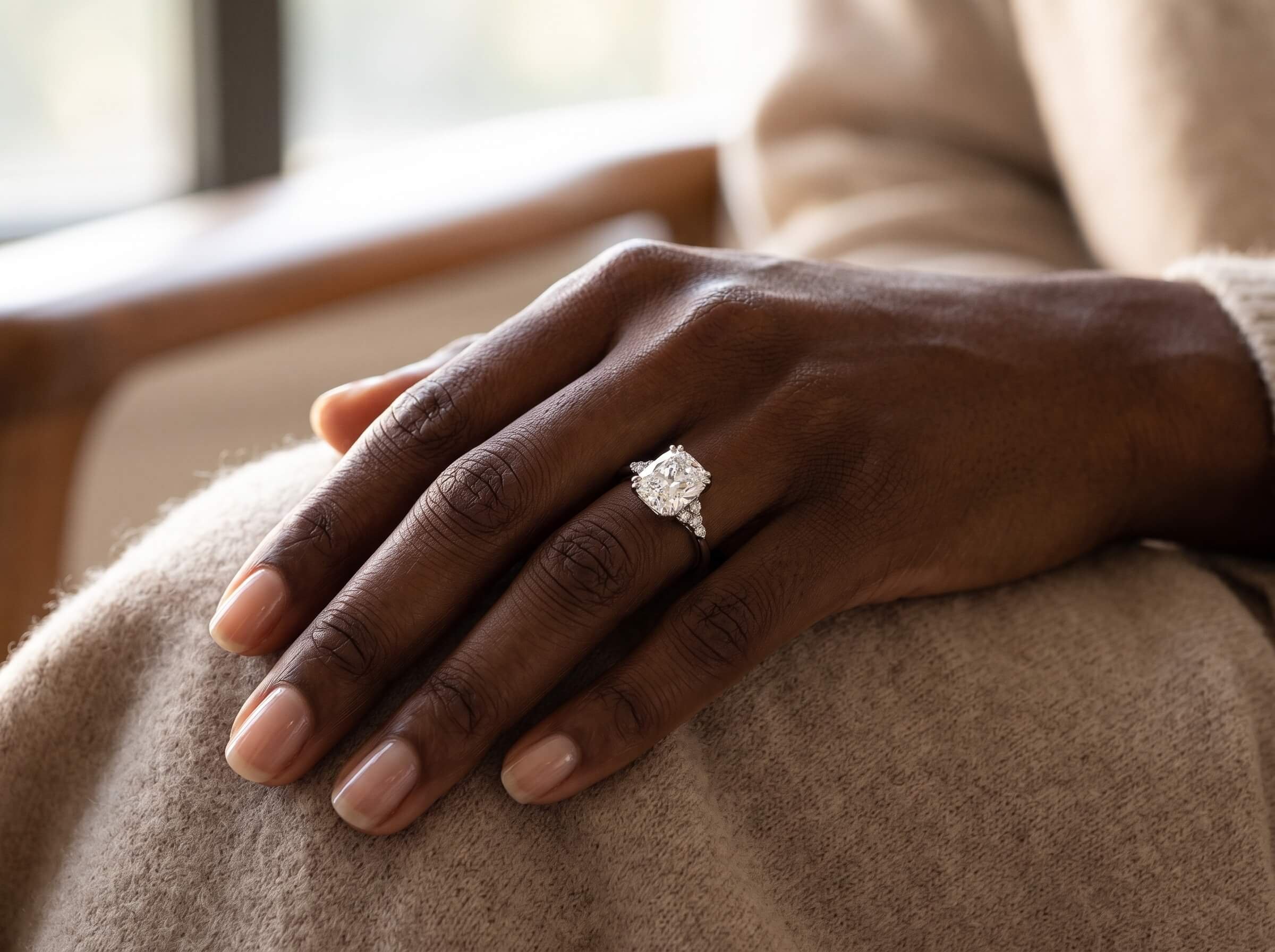 Close-up of a dark-skinned hand with manicured nails wearing a sparkling cushion-cut diamond engagement ring, resting on beige fabric.