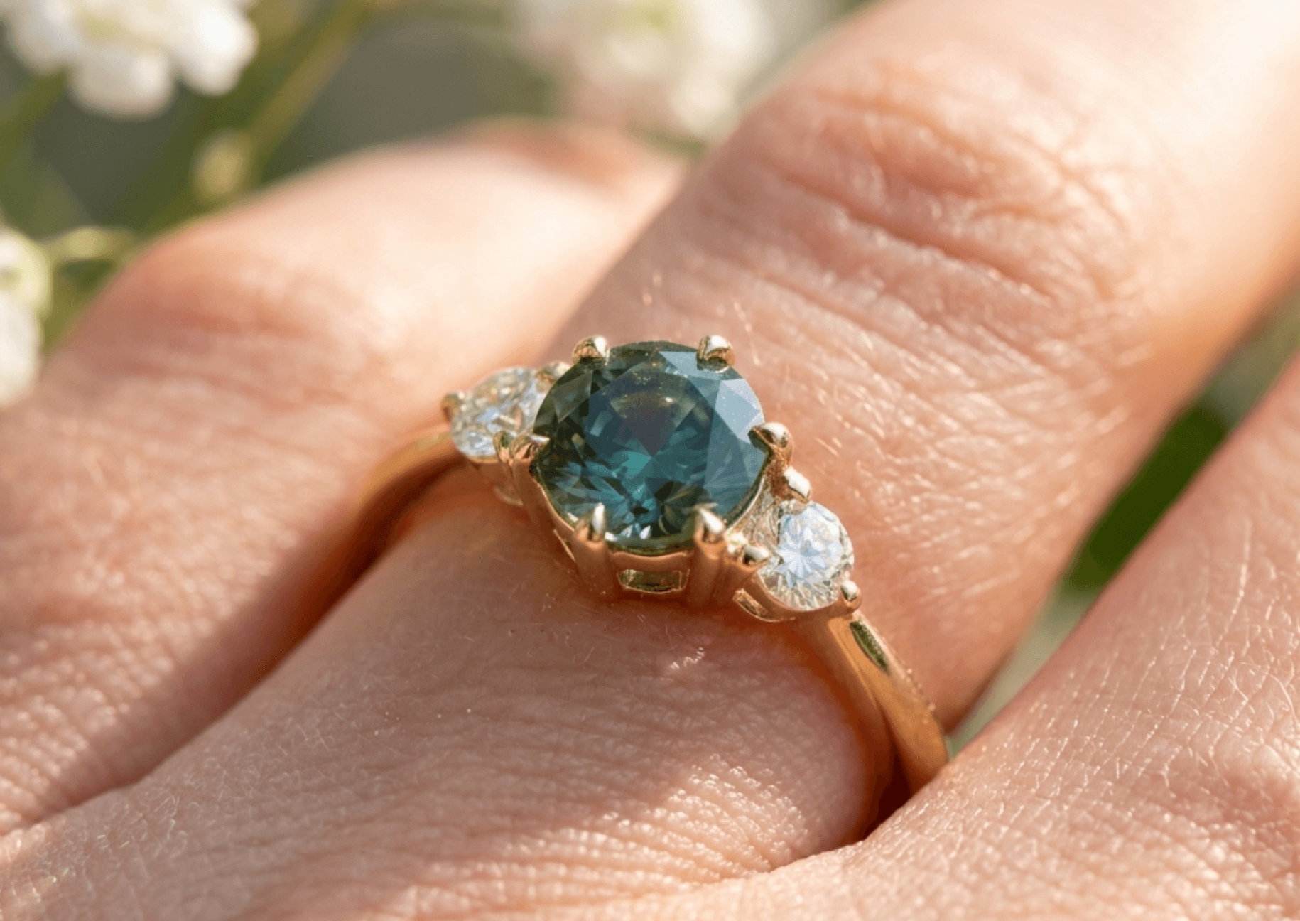 A close-up of a hand wearing a three-stone ring with a round teal sapphire center and two round diamond side stones on a yellow gold band, with soft white florals in the background.