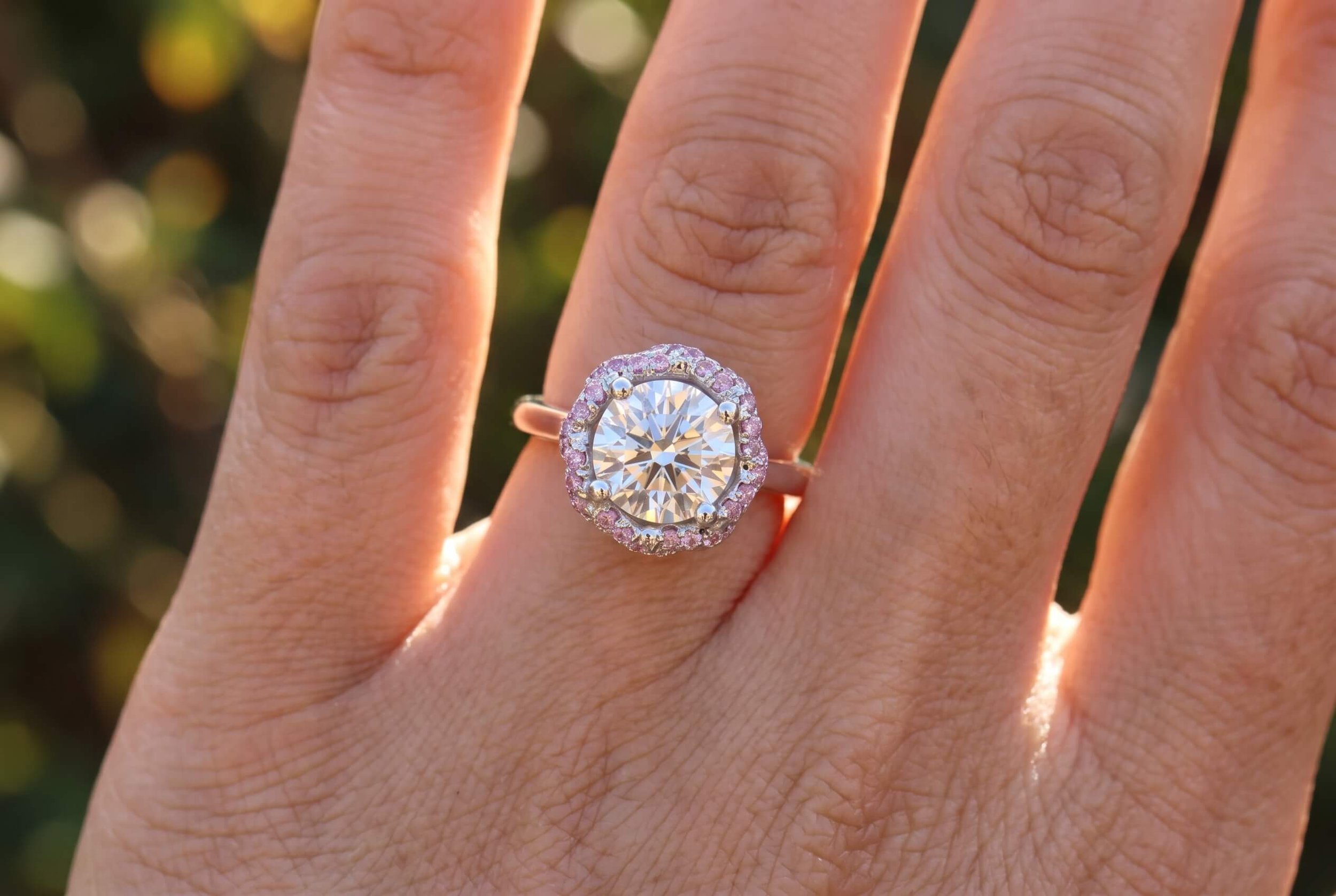 A close-up of a hand wearing a round brilliant diamond ring with a scalloped pink sapphire halo on a slim white gold band, in warm golden-hour light.