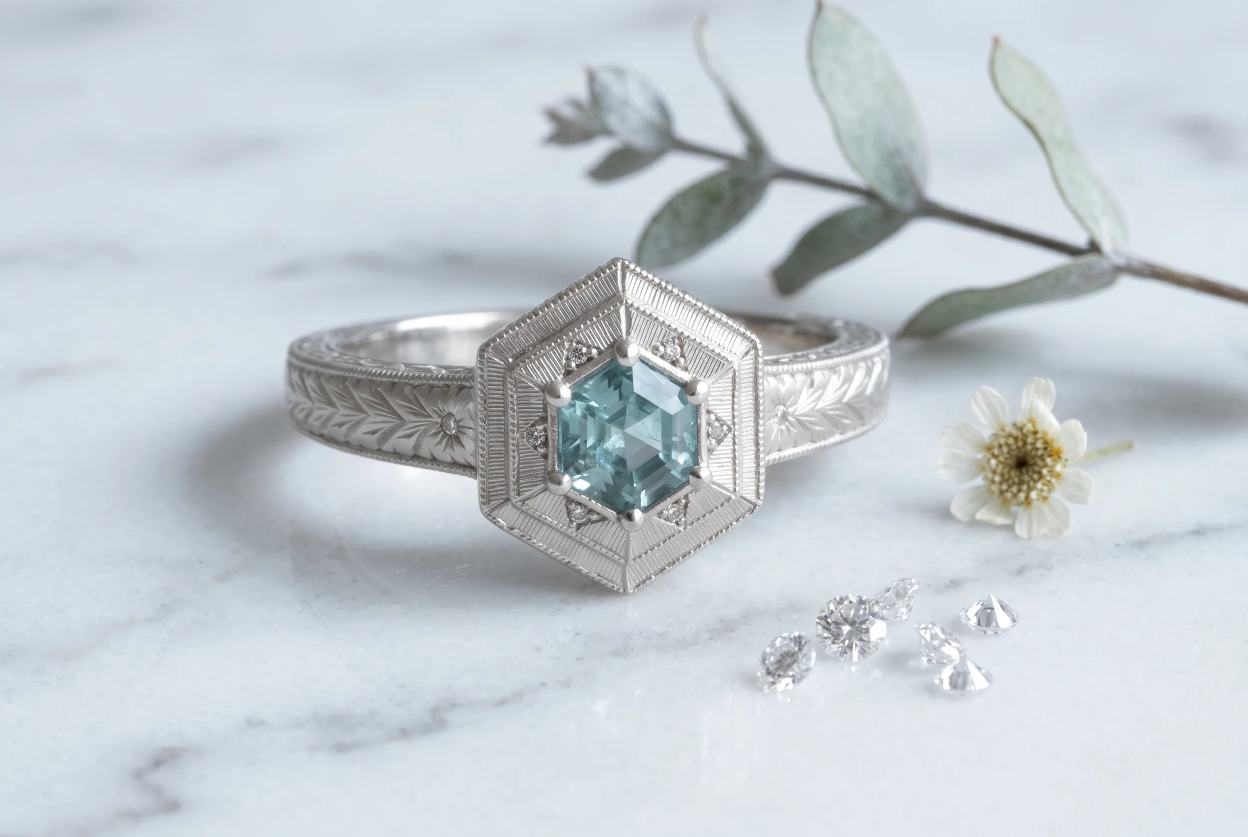 Hexagon teal sapphire engagement ring with engraved milgrain setting in white gold, styled on marble with loose diamonds and eucalyptus
