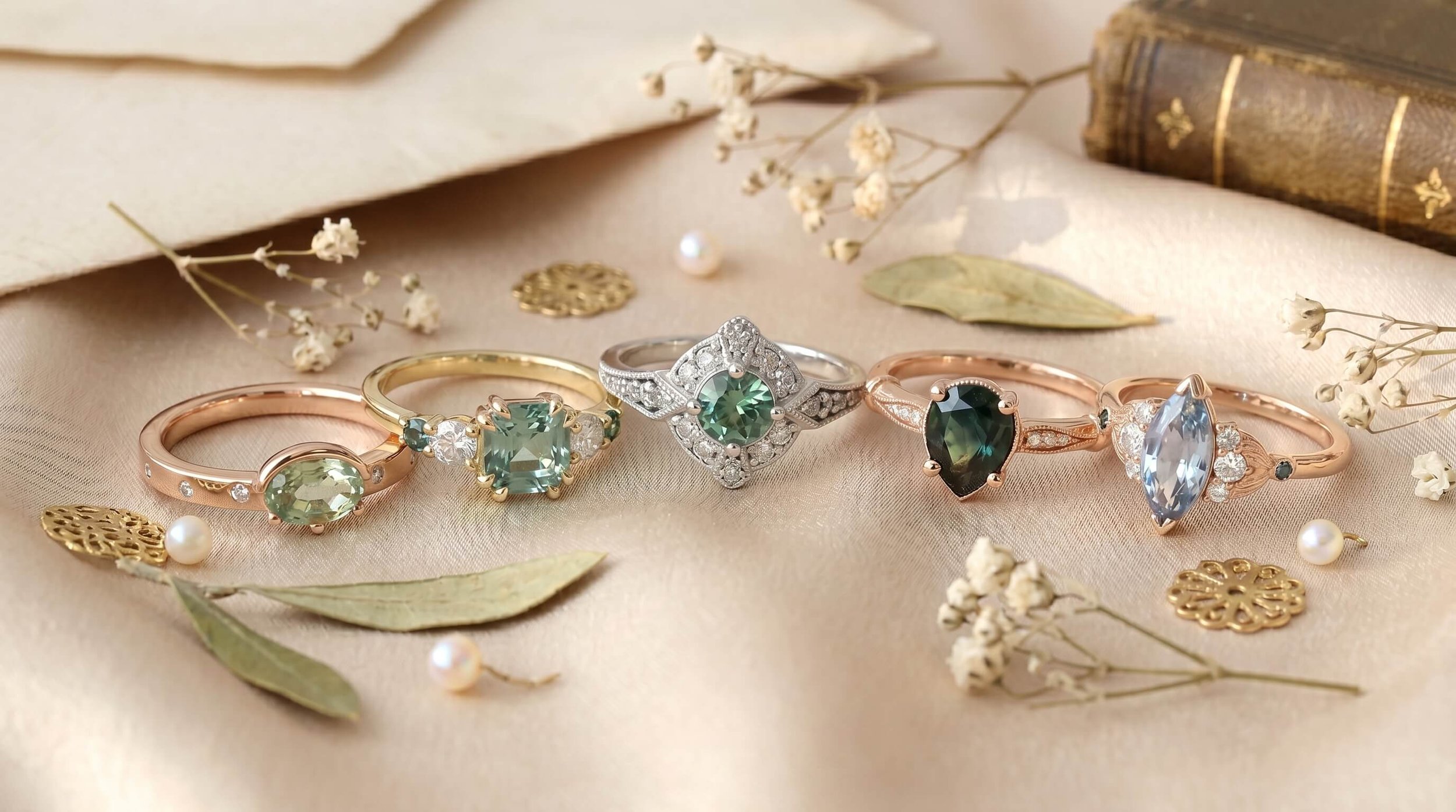 Collection of vintage-inspired green and blue sapphire engagement rings in rose, yellow, and white gold, styled on champagne satin with dried florals, pearls, and antique book