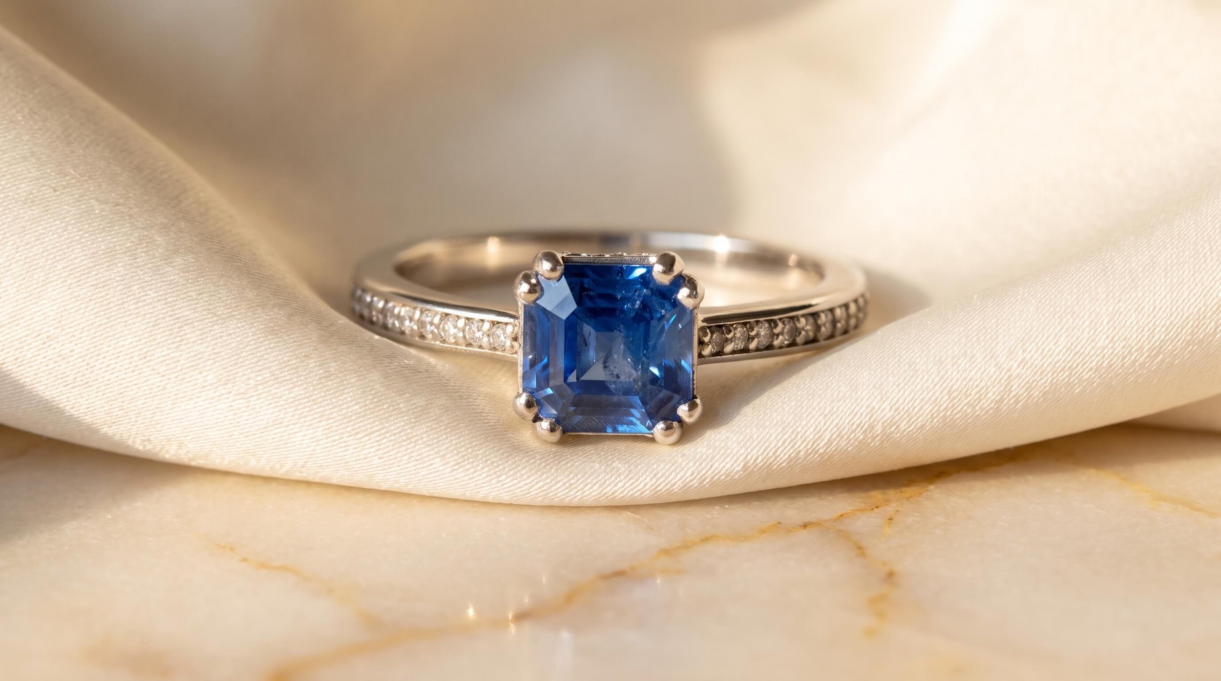 An Asscher-cut blue sapphire ring with double prongs and a diamond pavé band, displayed on draped ivory silk over marble.