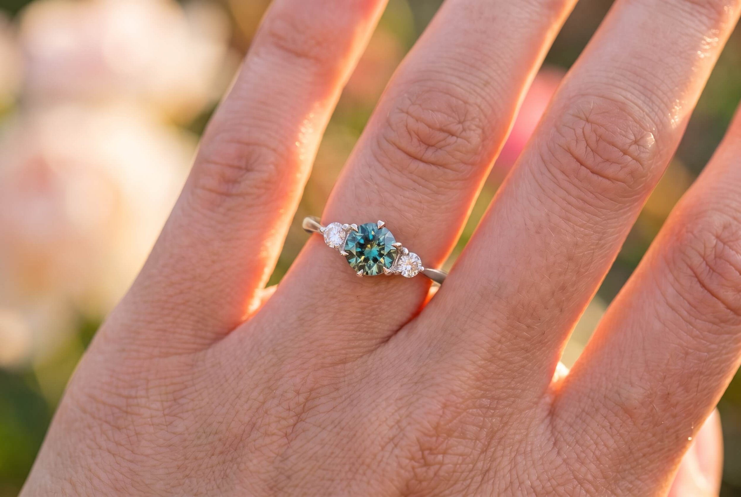 A close-up of a hand wearing a three-stone ring with a round teal-green center gemstone flanked by two round diamonds on a white gold band, in warm golden-hour light with soft floral bokeh.