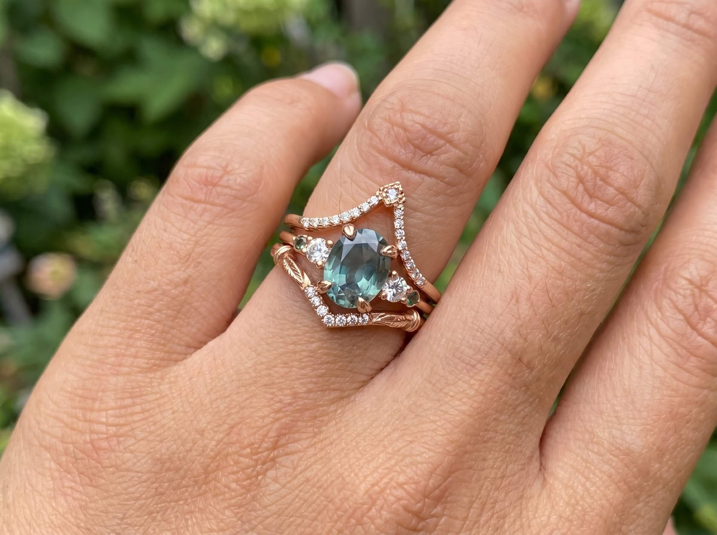 A close-up of a hand wearing a stacked ring set with an oval teal gemstone center ring and a crown-shaped diamond pavé band, set against a garden background.
