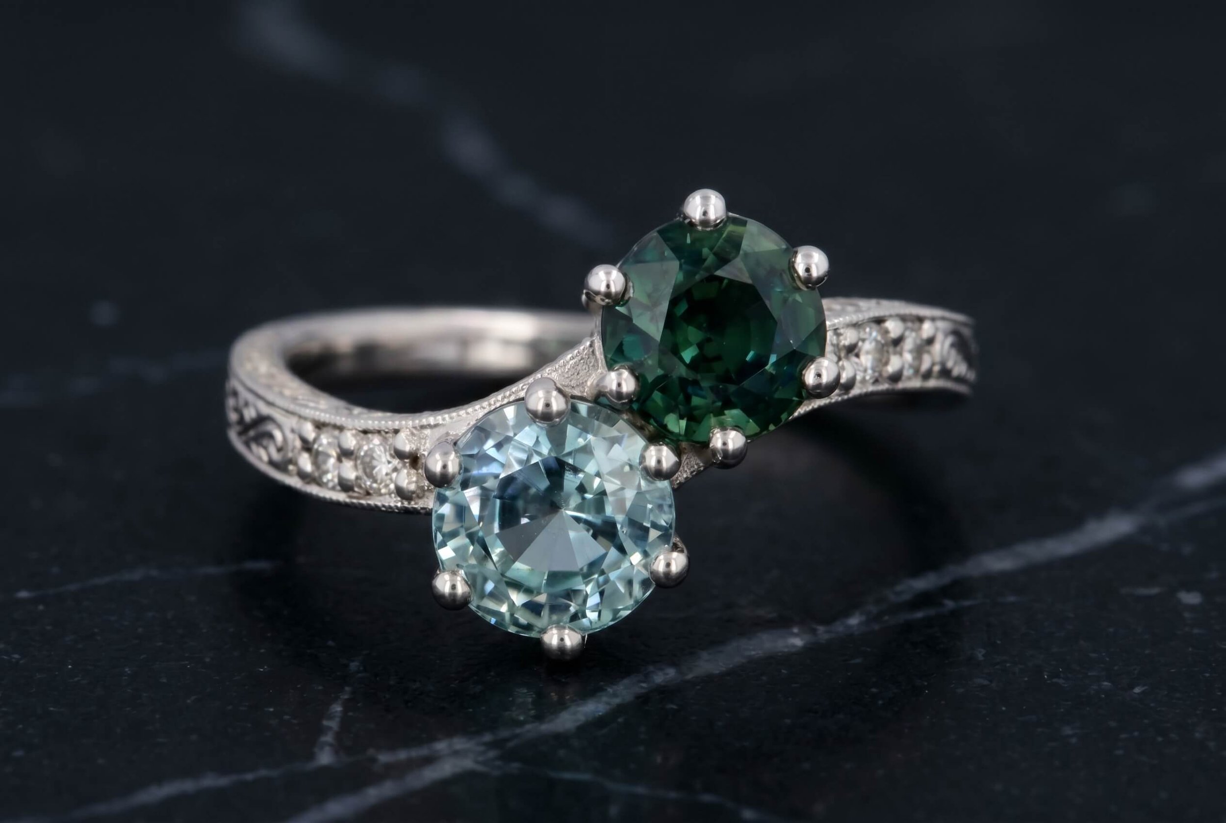 Two luxury rings on a dark marble surface: a silver ring with a large light blue aquamarine and another silver ring with a large deep green emerald, both featuring diamond accents.