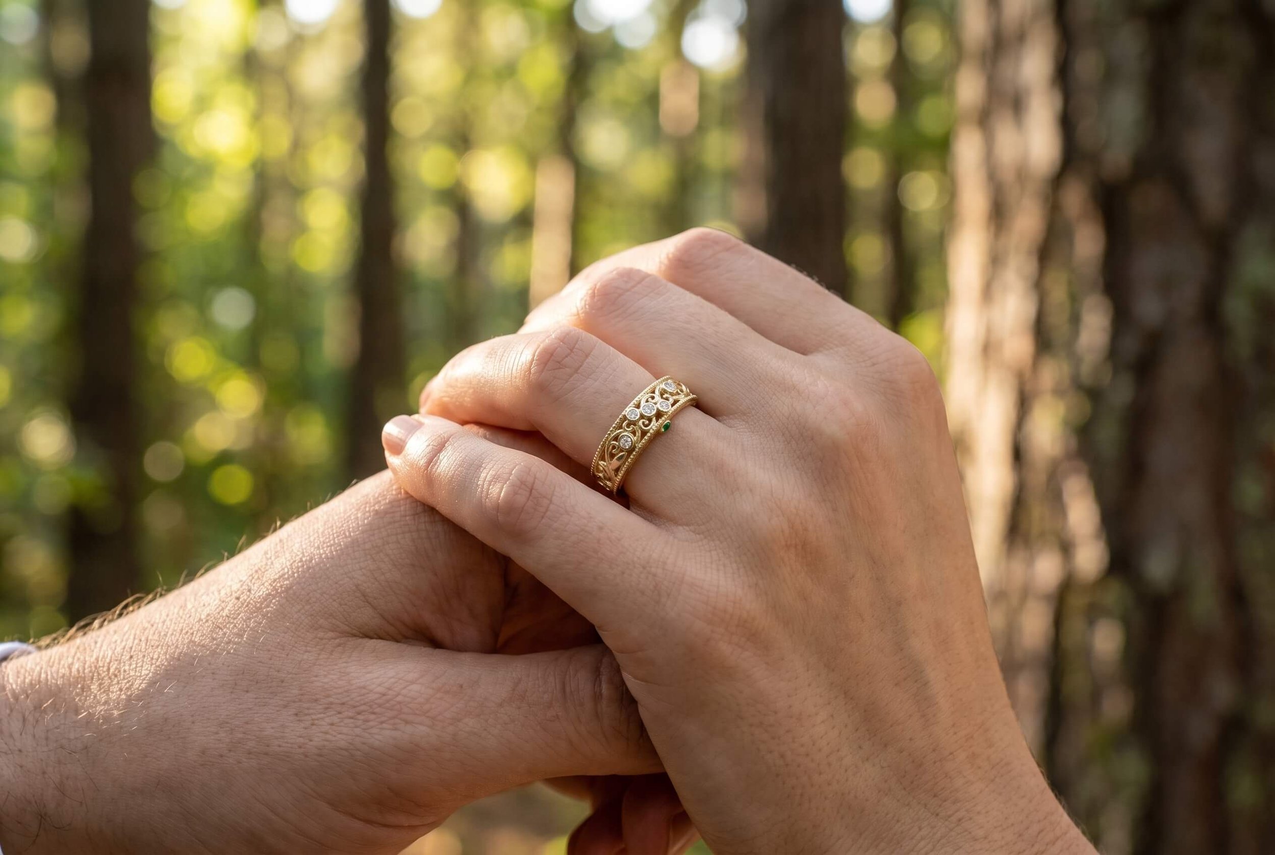 Two hands holding each other in a sunlit forest, with one hand wearing a wide yellow gold band featuring diamond bezels, filigree scrollwork, and a small green gemstone accent.
