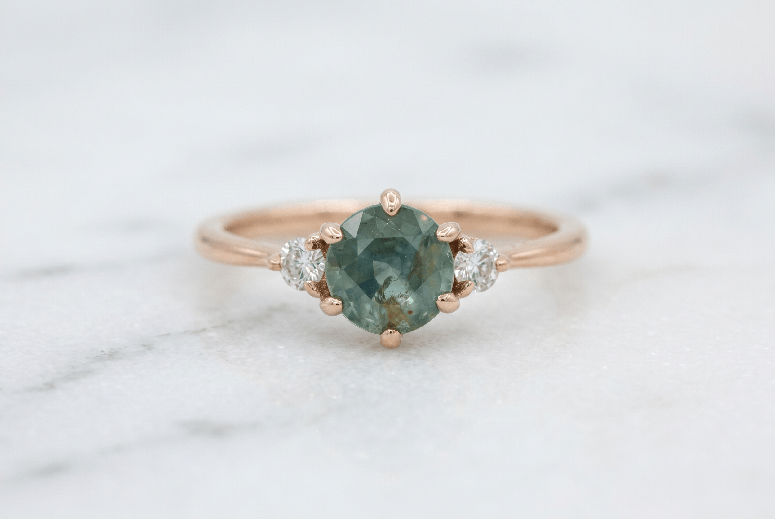 Round moss agate three-stone engagement ring with round diamond side stones in rose gold on white marble