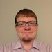 A young man with glasses and short hair, wearing a collared shirt, standing against a plain background.