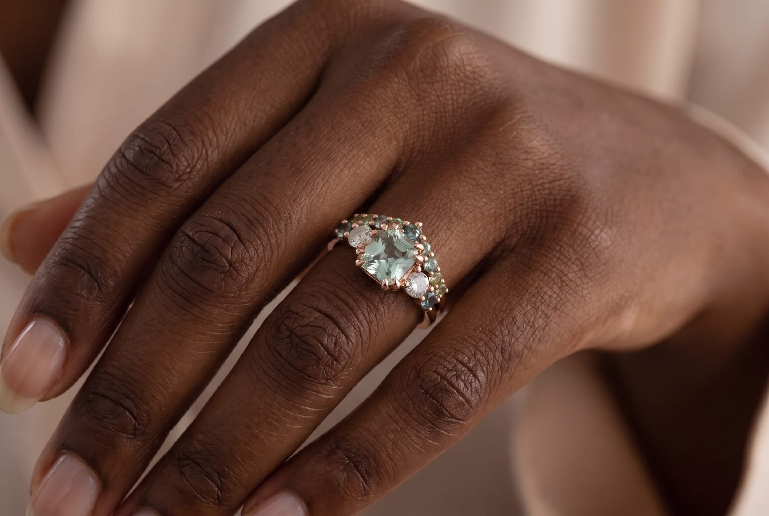 A close-up of a hand wearing a rose gold ring featuring a radiant-cut mint green gemstone surrounded by a cluster of teal, green, and white accent stones.