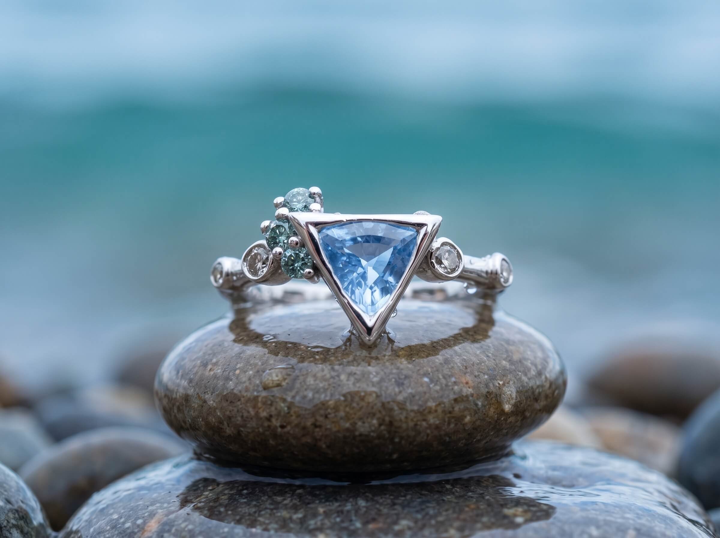 A white gold ring with a trillion-cut light blue gemstone, cluster of small green accent stones, and diamond side accents, balanced on a wet pebble with a blurred turquoise seascape behind it.