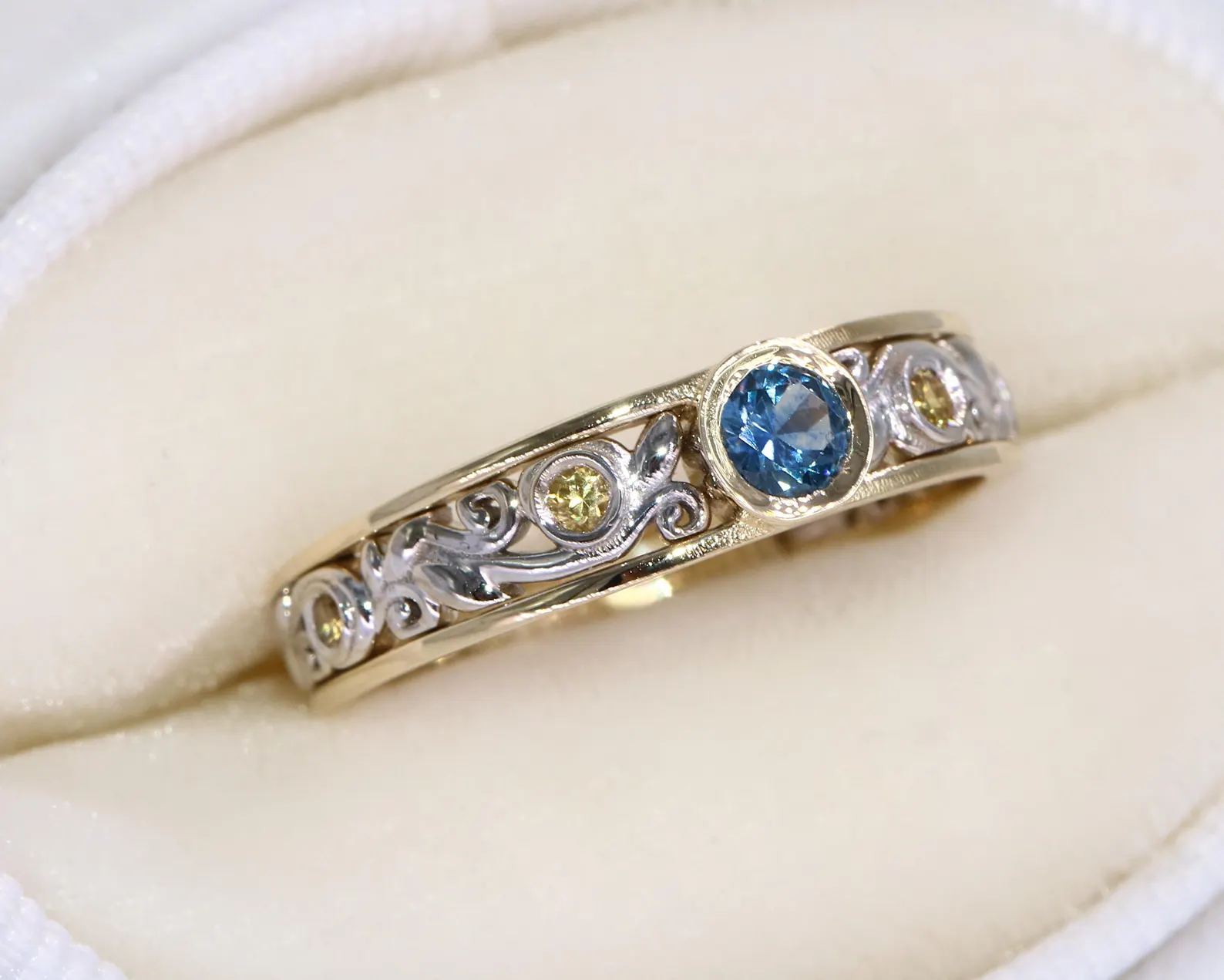 A gold and silver ring with intricate filigree designs, featuring a large blue gemstone in the center flanked by smaller yellow stones, placed on a soft padded surface.