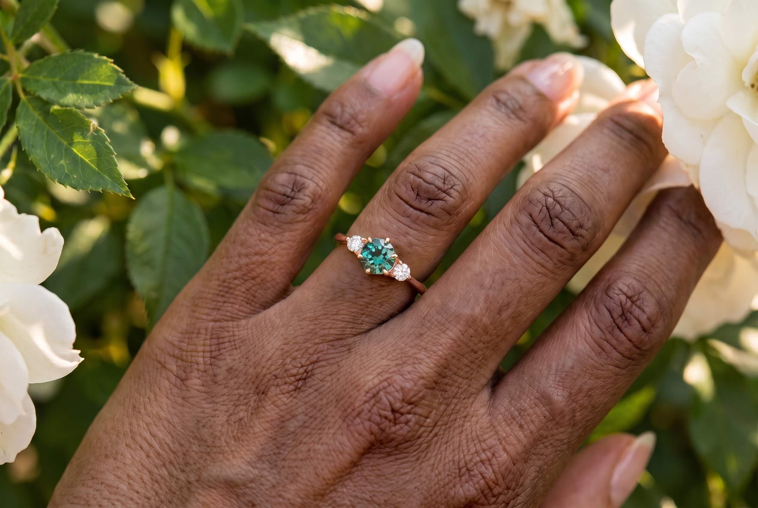 A close-up of a hand wearing a three-stone ring with a round teal-green center gemstone flanked by two round diamonds on a rose gold band, set against white roses and green leaves.