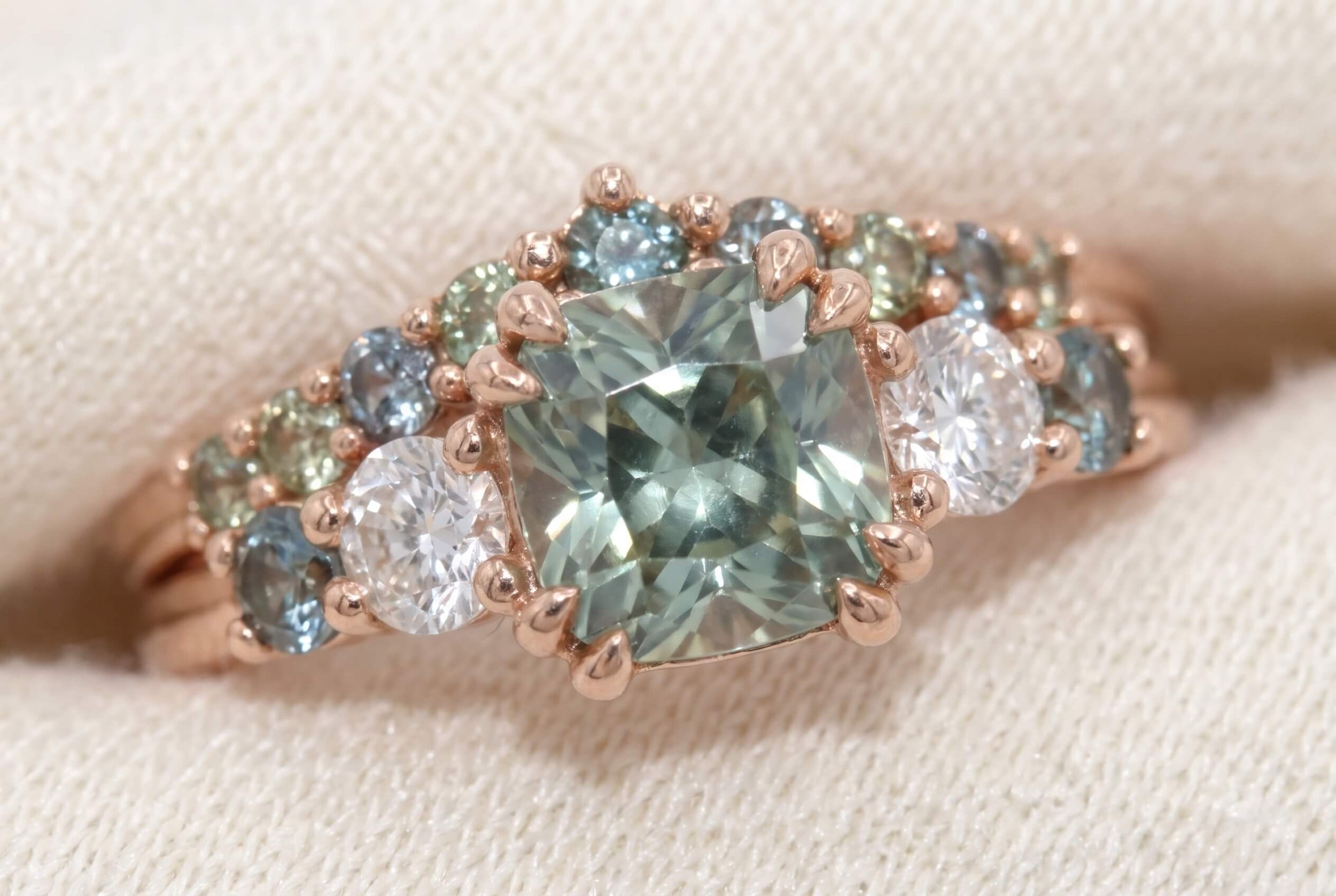 A macro close-up of a rose gold ring with a cushion-cut mint green center stone encircled by a mixed cluster of round diamonds and teal and green sapphires.