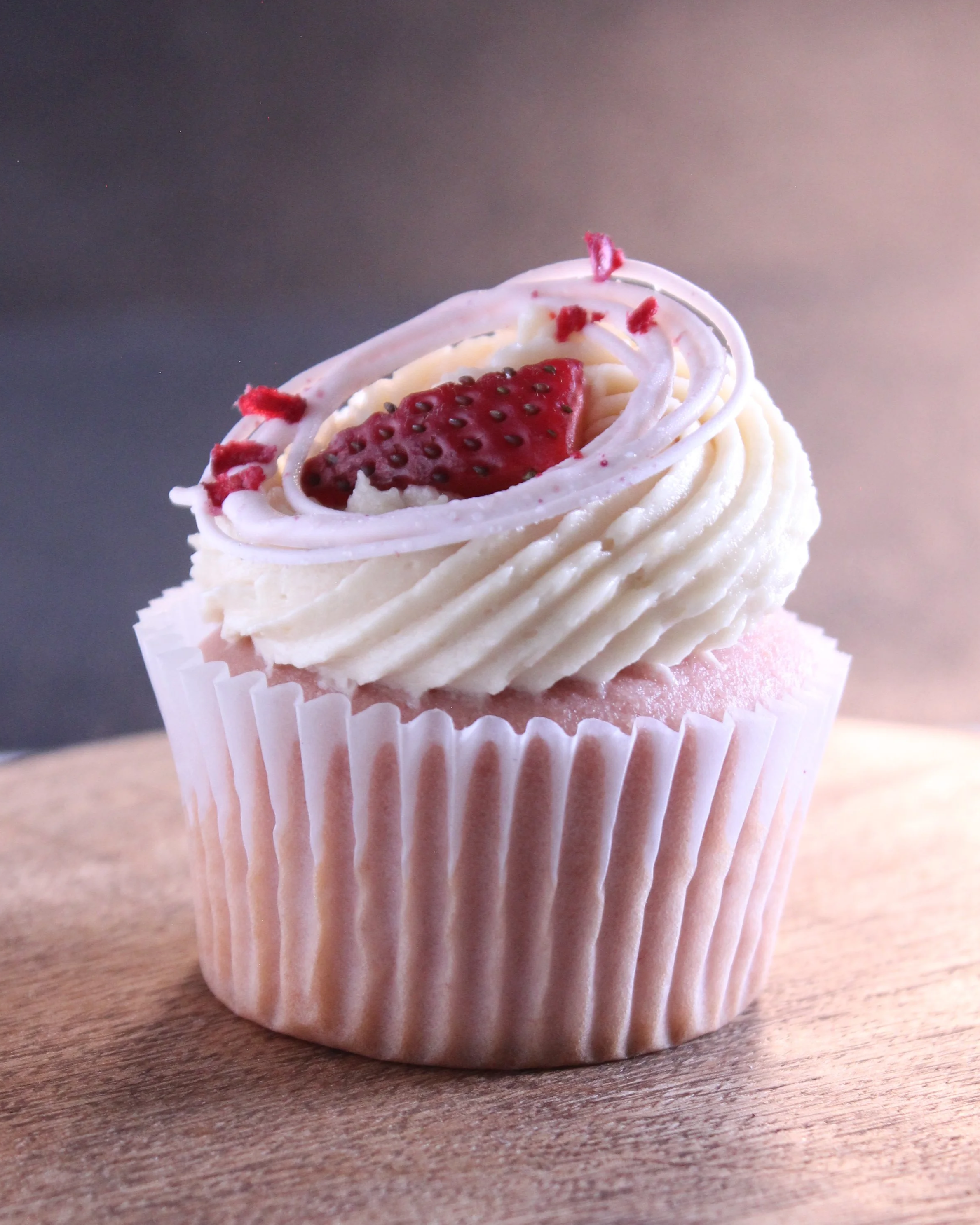 Pink cupcake with white frosting, topped with strawberry slice and red sprinkles, on a wooden surface with a blurred background.
