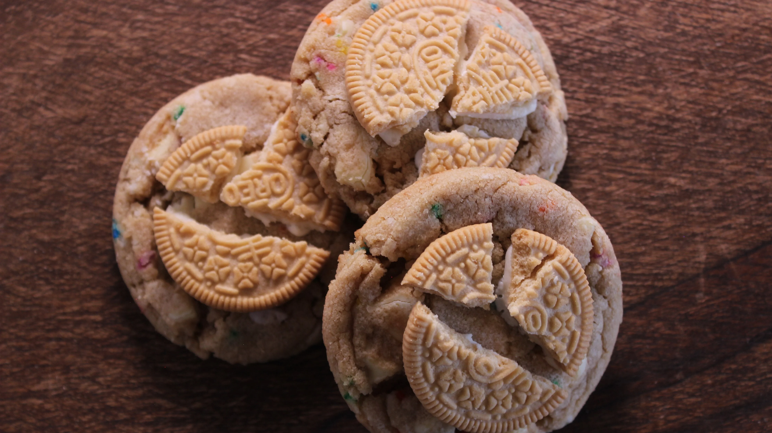 Three cookies on a wooden surface, two with colorful sprinkles and broken pieces of cookies on top, one with half of a miniature Reese's peanut butter cup embedded on top.