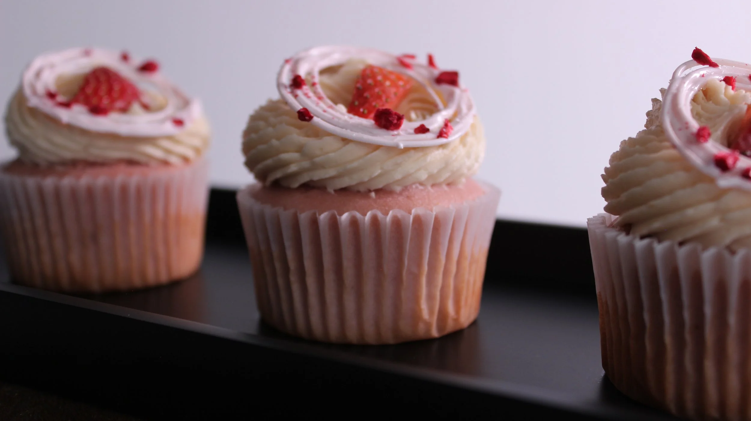 Three pink cupcakes topped with swirled white frosting, strawberries, and red decorative sprinkles on a black rectangular tray.