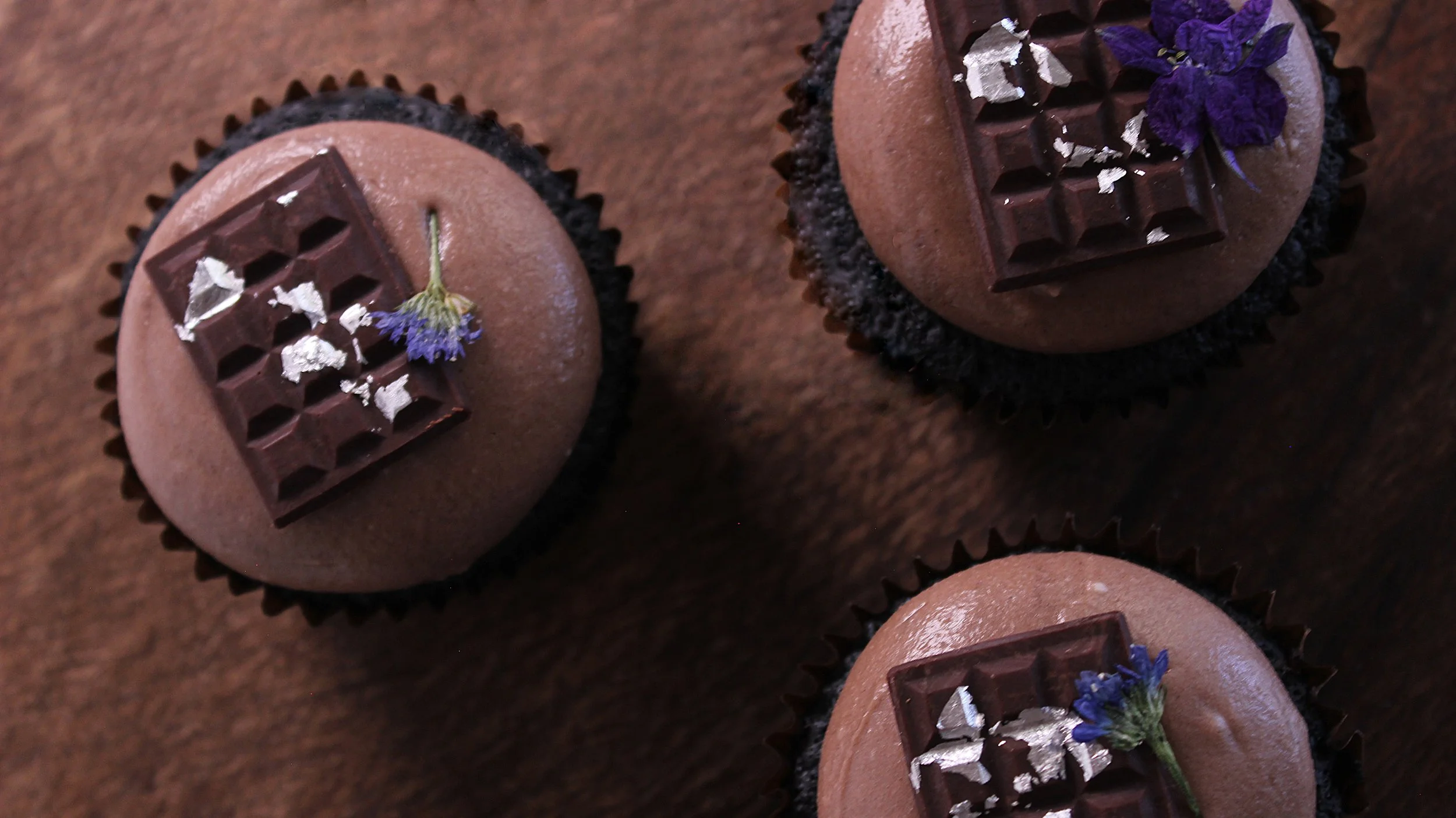 Three chocolate cupcakes topped with a square of dark chocolate, silver leaves, and small purple flowers, on a wooden surface.