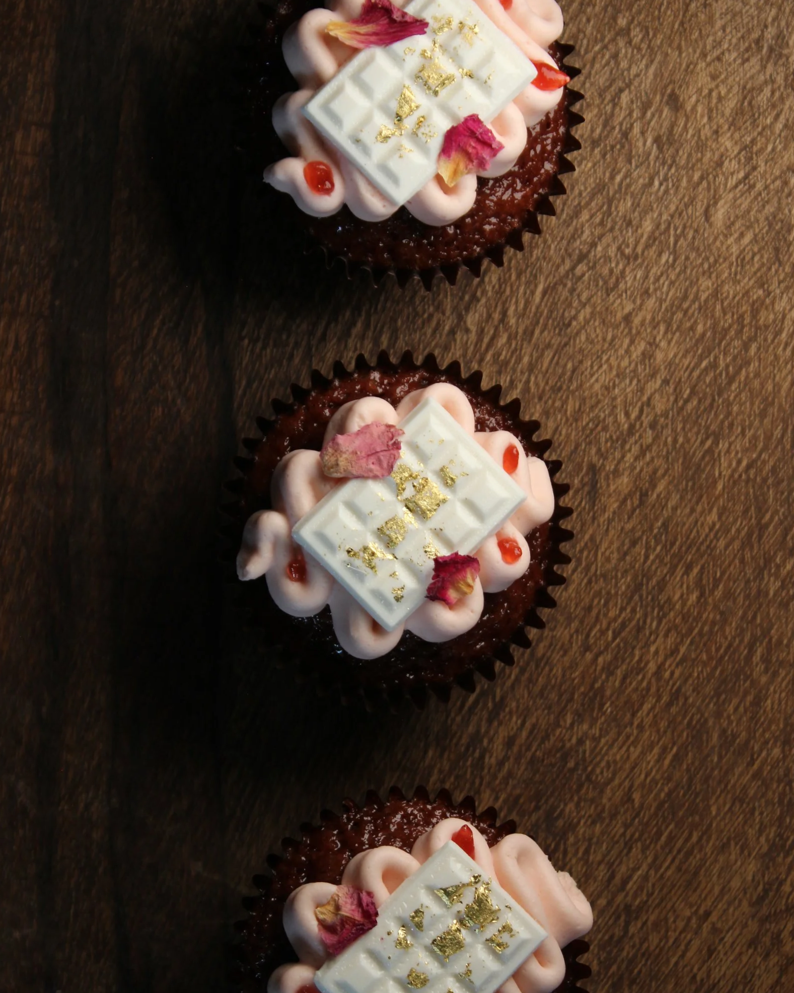 Three cupcakes with pink frosting, white chocolate squares with gold flakes, small red and pink decoration, and dried pink rose petals, arranged in a vertical line on a wooden surface.