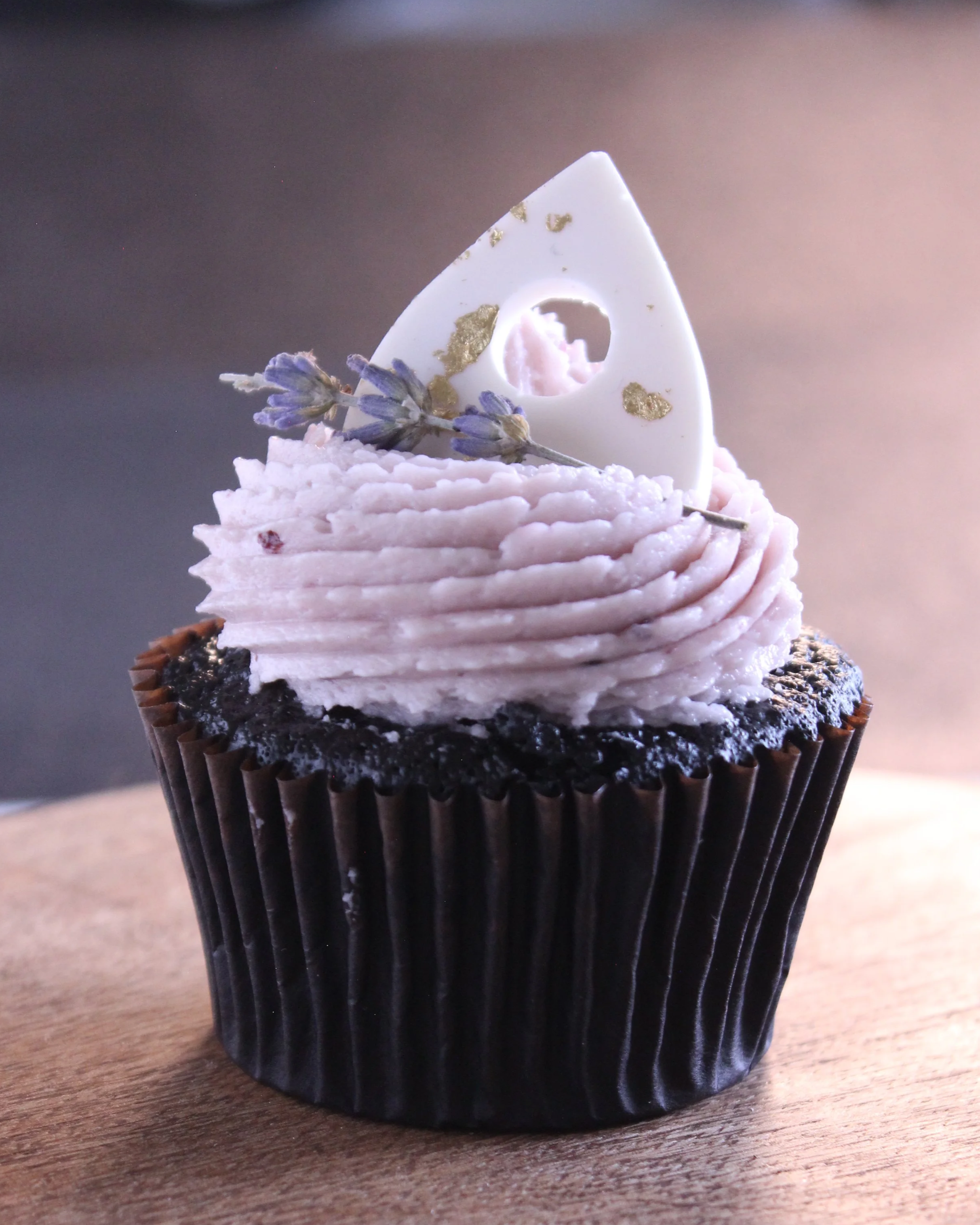 Chocolate cupcake with pink frosting, topped with a white decorative triangle, a sprig of lavender, and gold accents.