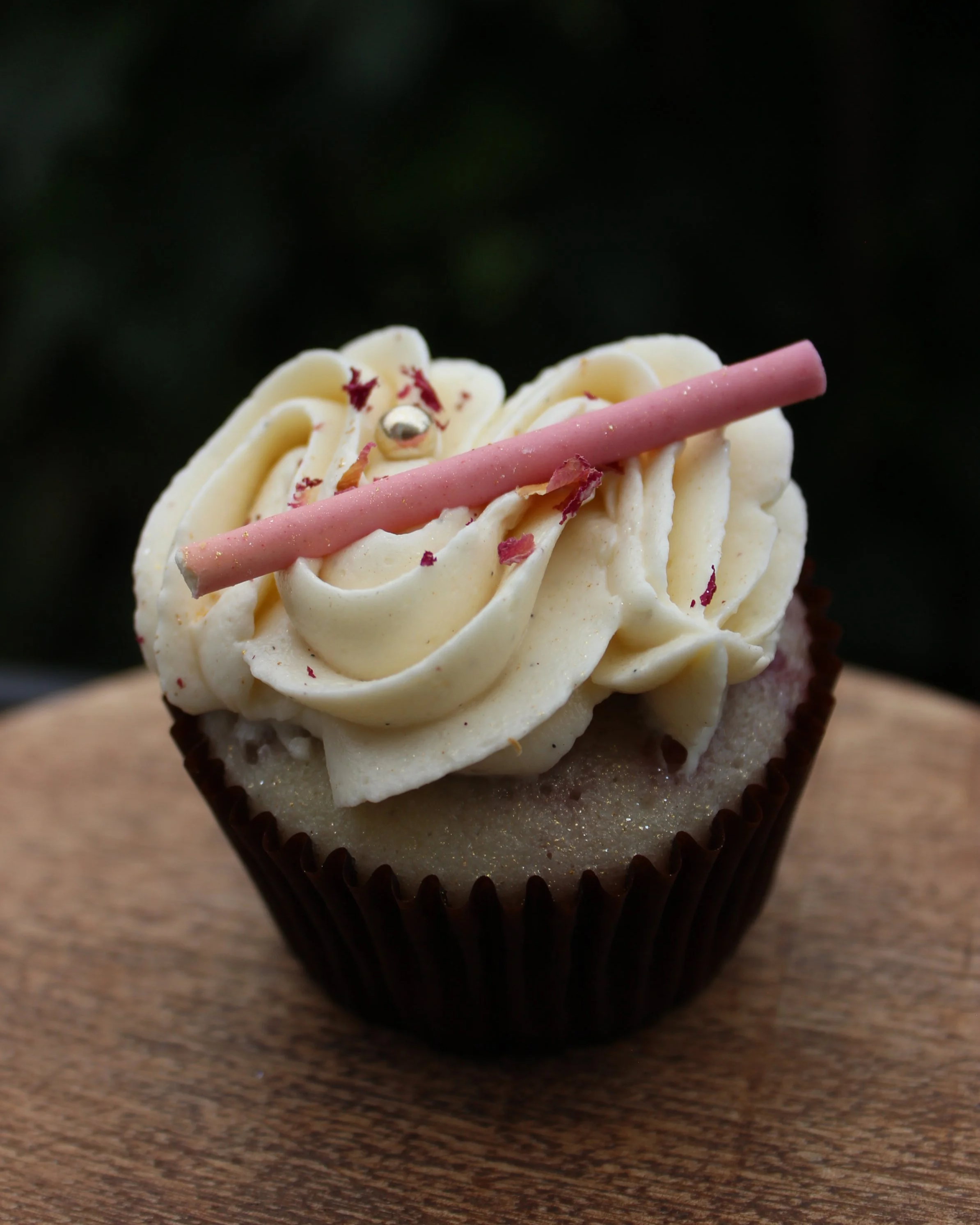 A cupcake with white frosting, a pink stick, and a silver ball decoration, sitting on a wooden surface.