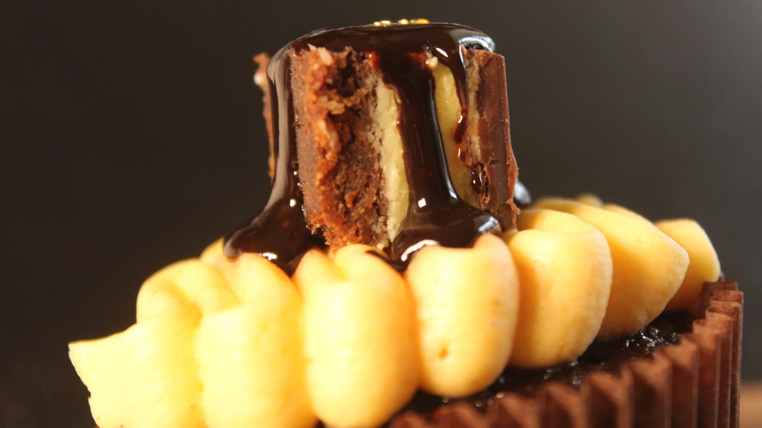 A close-up of a piece of chocolate cake topped with chocolate ganache, with a yellow piped frosting decoration on a chocolate base.