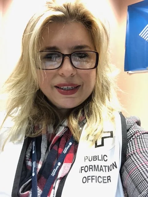 A woman with blonde hair wearing glasses and a checked shirt, taking a selfie in front of a purple wall. She is wearing a white vest with the words 'Public Information Officer' printed on it.