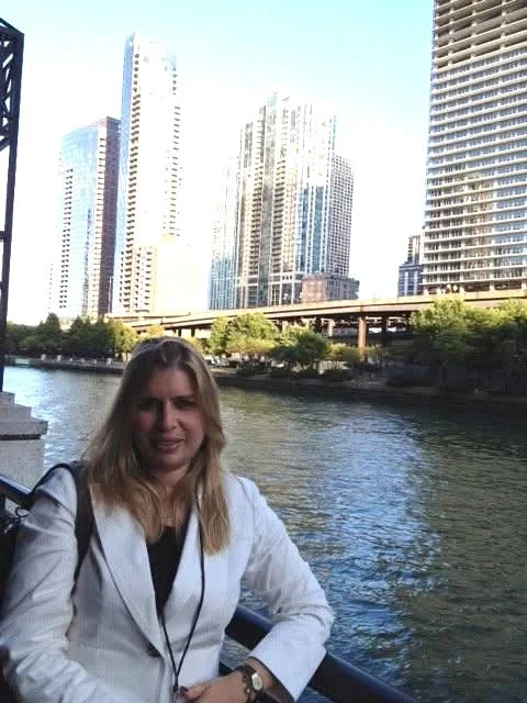 Woman with blonde hair wearing a white blazer standing near a river with city skyscrapers in the background.