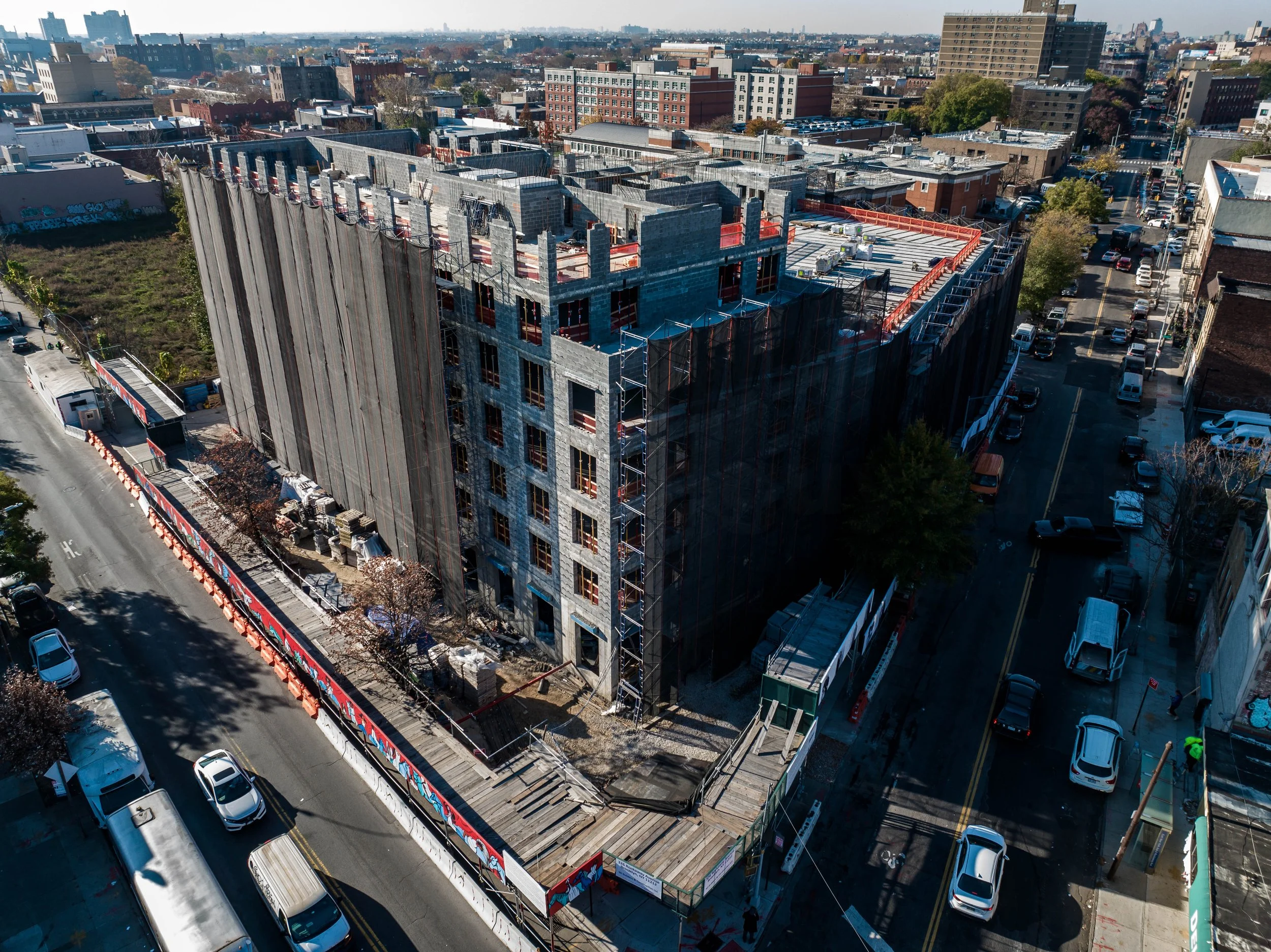 An aerial view of a building under construction on a city street, with construction scaffolding and barriers, surrounded by parked cars and other city buildings in the background.