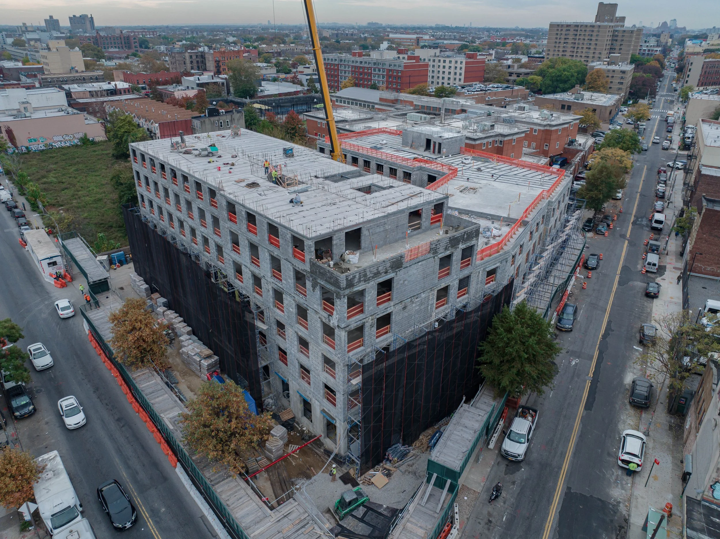 Aerial view of a multi-story building under construction in an urban area, with construction workers on the roof, scaffolding along the sides, and a crane in the background. Surrounding streets are filled with parked cars and nearby buildings.