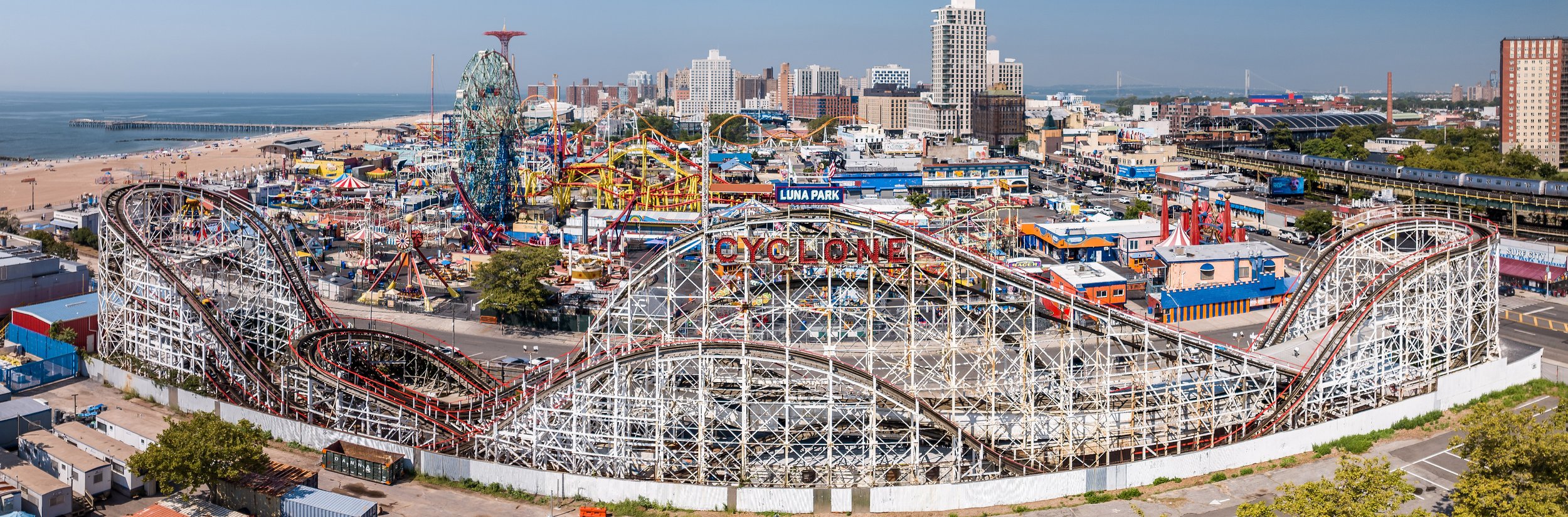 A large amusement park in Brooklyn near a beach with a roller coaster named 'Coney Island Cyclone', various other rides, attractions, and taller city buildings in the background.