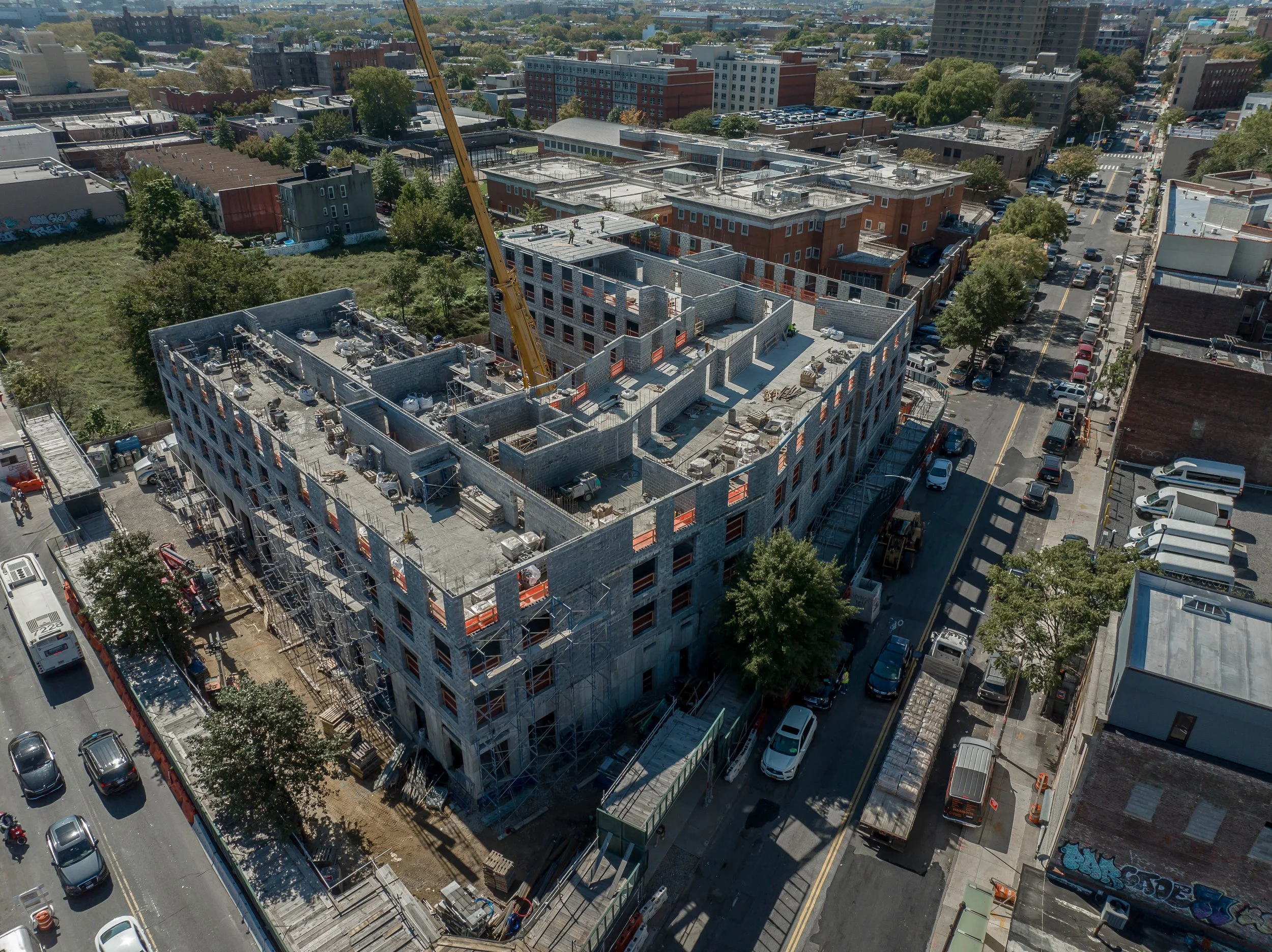Aerial view of a city construction site with a partially built multi-story building, construction crane, scaffolding, vehicles, trees, and surrounding city streets