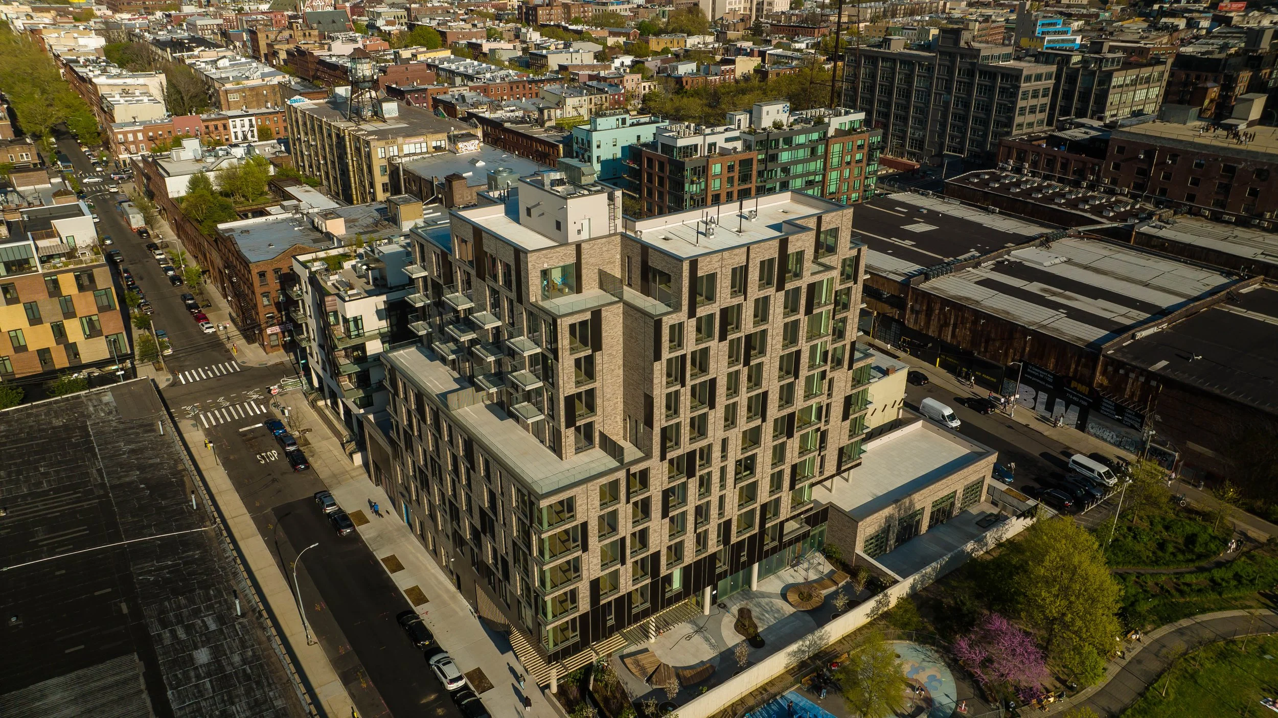 Aerial view of a modern multi-story residential building with multiple balconies, surrounded by other urban buildings and streets with parked cars, trees, and a park with a playground.