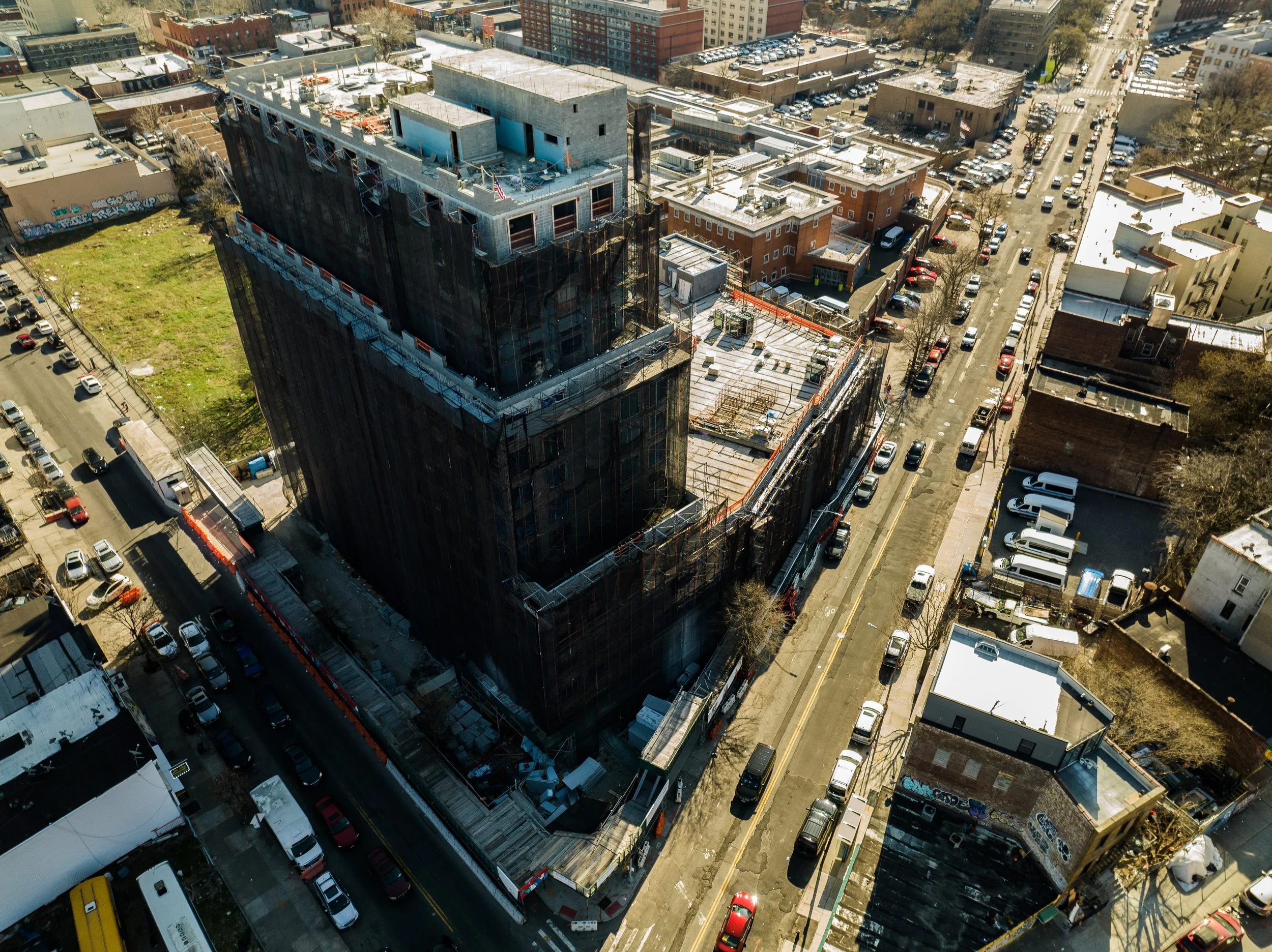 Aerial view of a construction site with a tall building under construction, surrounded by parked cars and nearby buildings on a city street.