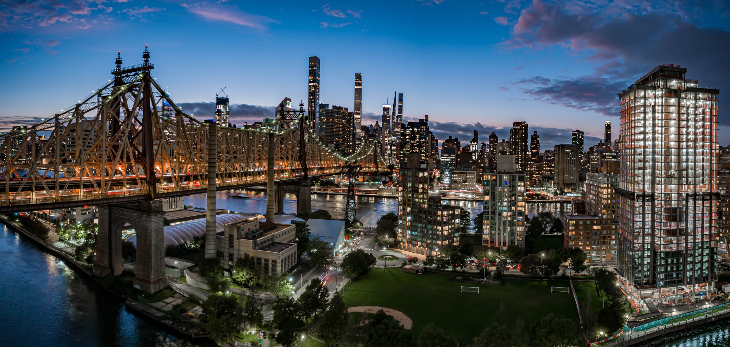 Nighttime cityscape of New York City featuring the 59th Street Bridge and Roosevelt Island over the East River with illuminated skyscrapers and city lights.