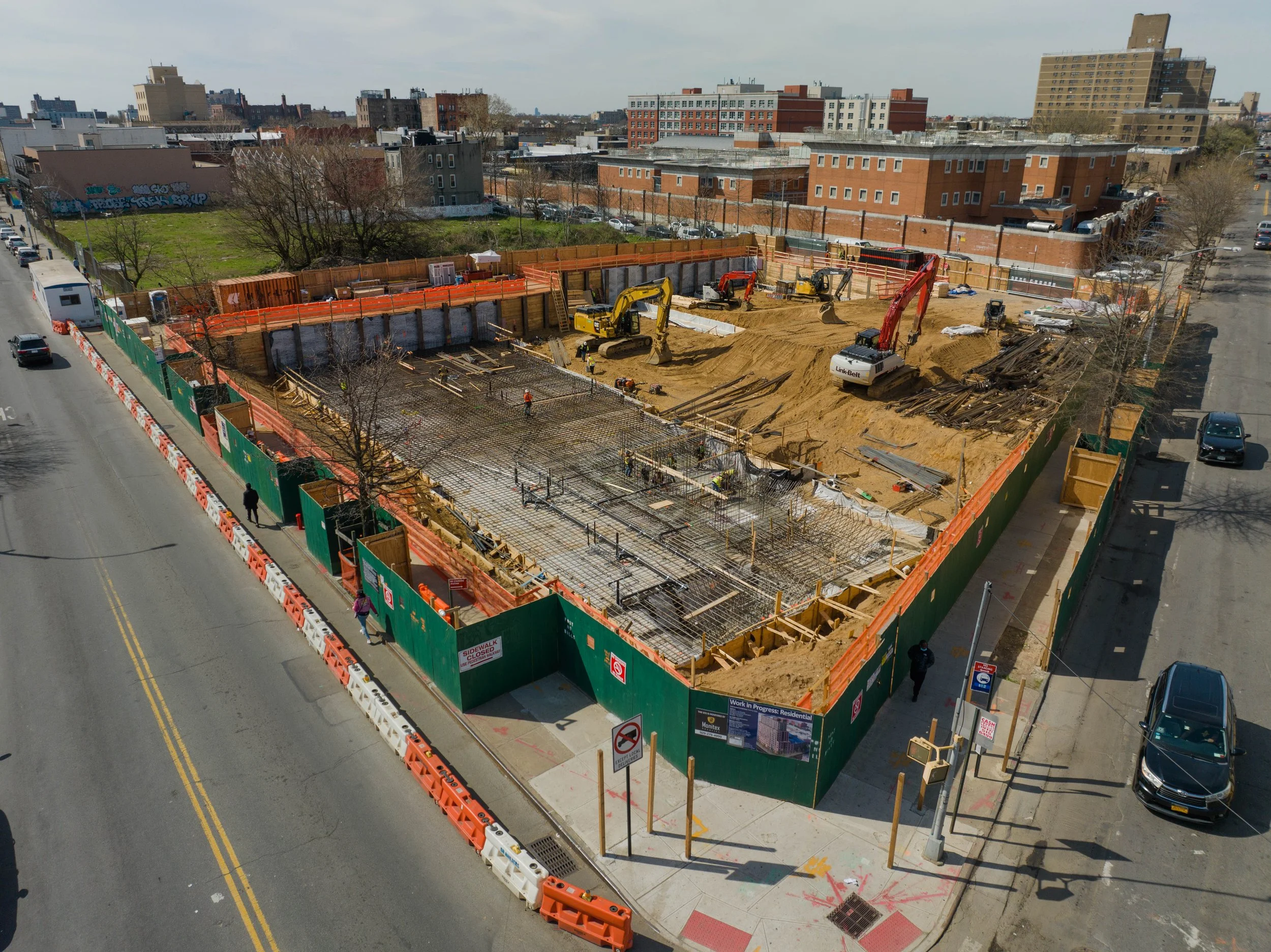 A construction site surrounded by green and orange safety barriers, with multiple workers and heavy equipment including excavators and concrete pouring in progress. Streets run alongside with some pedestrians and parked cars.