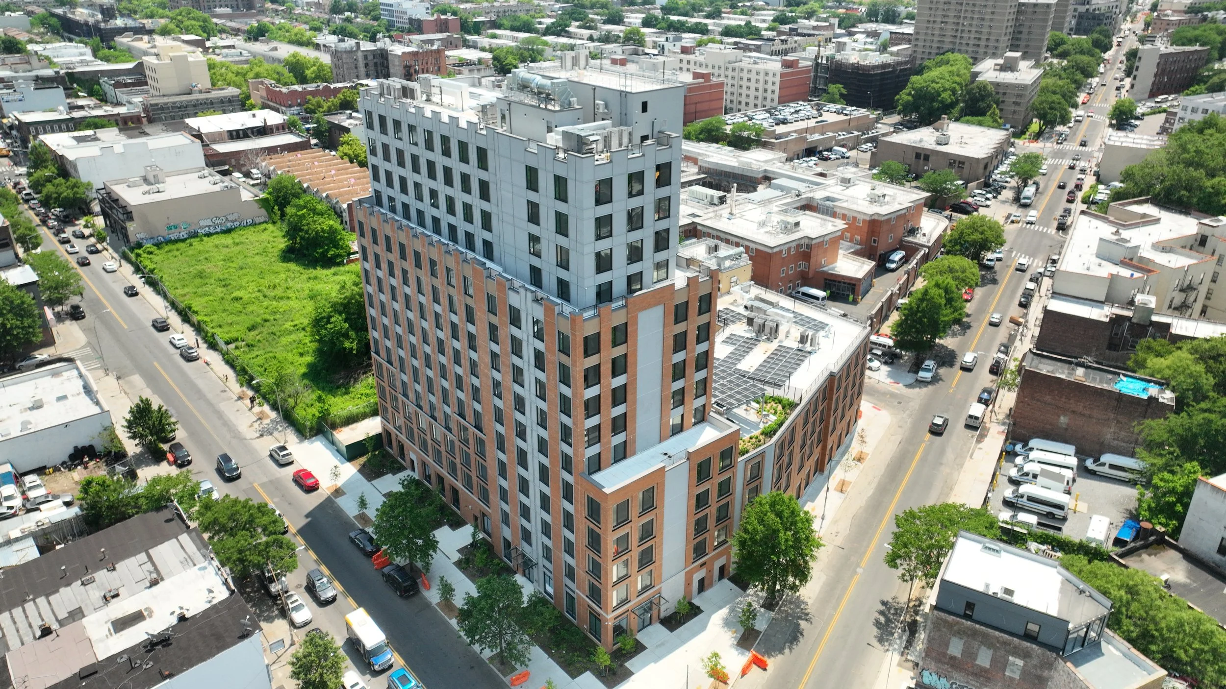 Aerial view of a modern high-rise building in a city neighborhood, surrounded by streets, cars, and lower buildings with trees and a green vacant lot nearby.