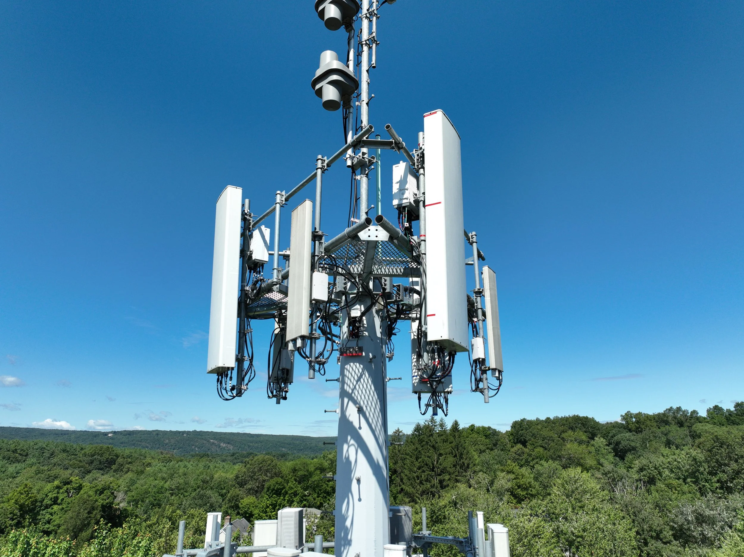 A close-up of a telecommunication tower with various antennas and dishes mounted on a metal structure, set against a clear blue sky with a green, forested landscape in the background.