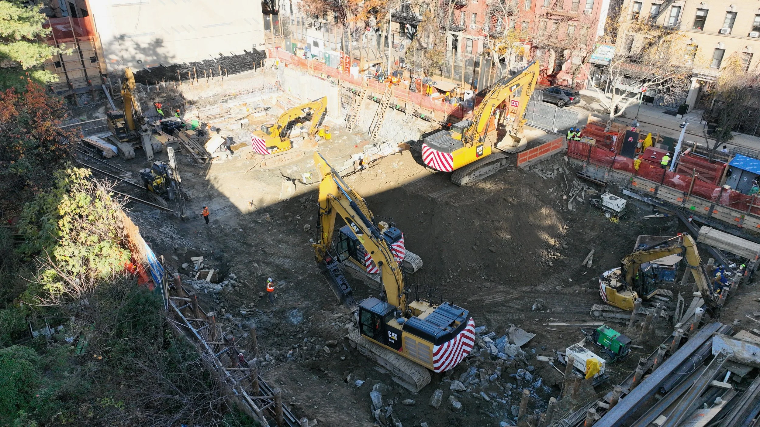Construction site with multiple excavators working on digging and moving earth, surrounded by safety barriers and workers in safety vests, in an urban area with buildings and trees.