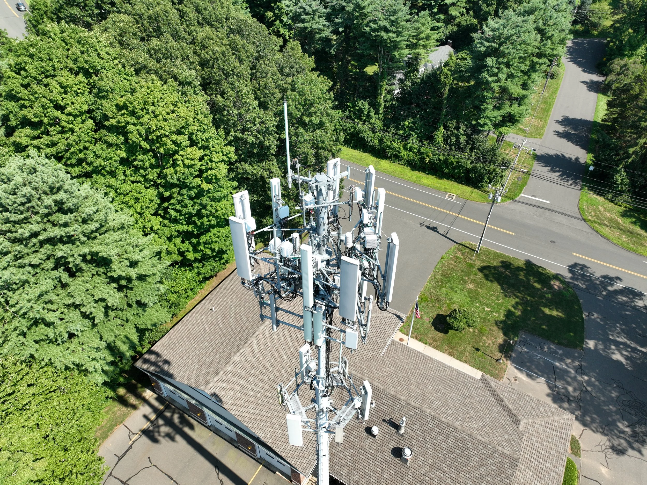 A tall communication tower equipped with multiple antennas and electronic devices, situated on a brick building surrounded by lush green trees and intersecting roads.